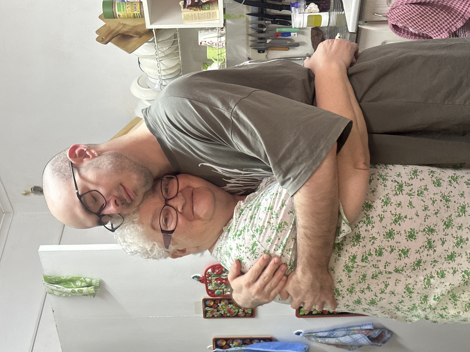 This indoor photograph, taken in a kitchen in Aktanysh, Russia, captures two individuals in a close embrace. An older woman with white, curly hair and reddish-brown framed eyeglasses occupies the left side of the frame, smiling towards the viewer. She wears a white top adorned with a green floral pattern. A younger bald man, wearing black-framed glasses and a silver ear cuff, stands to her right, with his eyes closed and his head gently resting against the woman's head and shoulder. He is dressed in a dark olive green oversized t-shirt featuring a subtle light graphic. Their arms are interlocked around each other's torsos in an affectionate gesture. The background reveals a domestic kitchen setting with white walls. On the left wall, multiple decorative potholders with folk-art style floral and berry designs are hung, alongside a blue and a plaid towel. The right side of the scene features a white wire dish rack filled with clean white plates and bowls. Above this, a white shelving unit holds kitchen items, including a green spice jar, a box labeled "Кофе" (Coffee), and several mugs. Below the shelf, a magnetic strip displays a collection of kitchen knives. A pink and white checkered towel hangs on a hook near the visible portion of a sink.