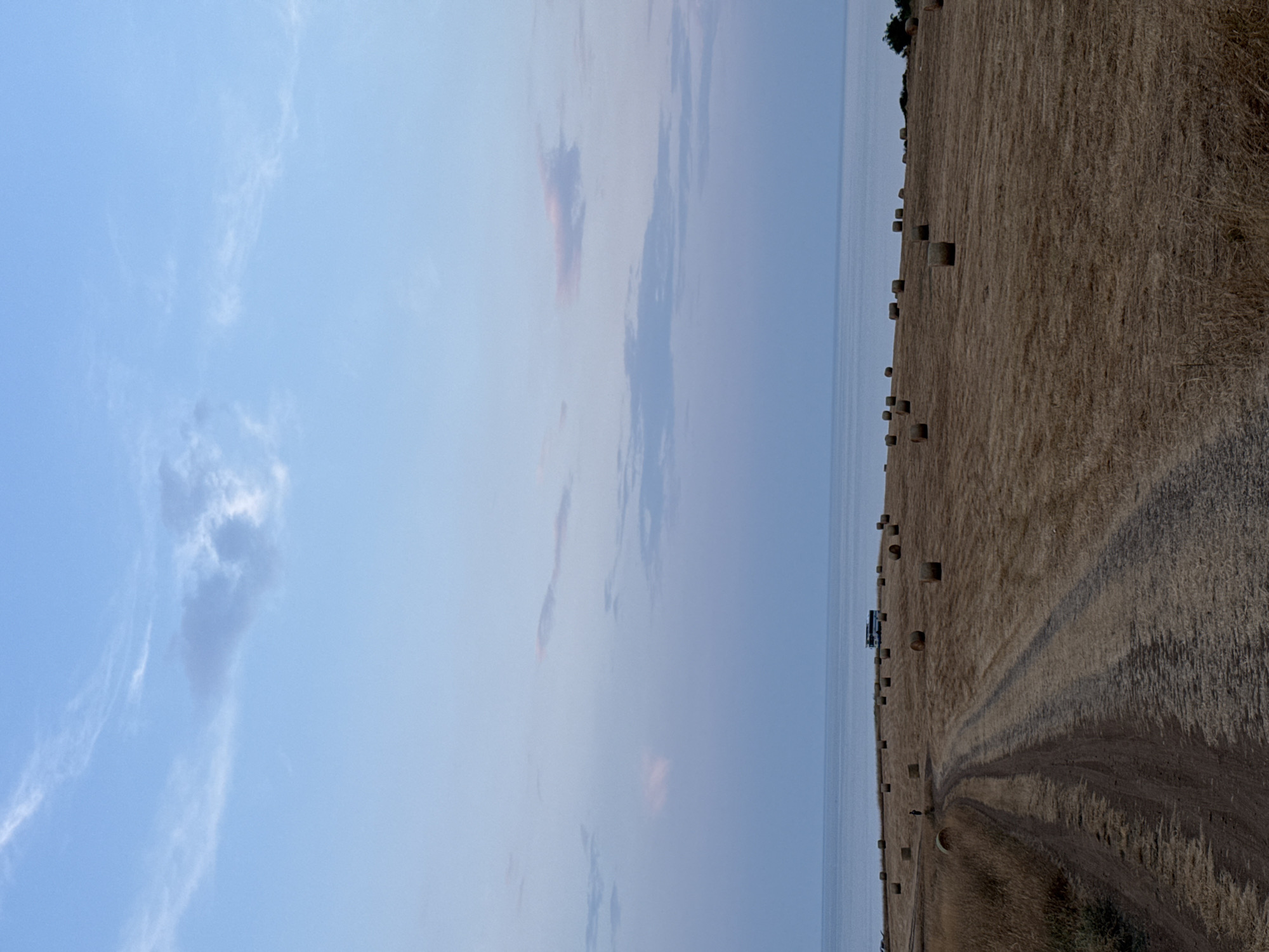 A dirt road cuts through a harvested field dotted with numerous round hay bales, leading towards a calm sea under a pale blue sky with wispy clouds. A small, dark-colored figure stands near a hay bale on the left side of the road. In the distance, on the horizon line where the field meets the sea, a white bus is parked among the hay bales. The overall scene suggests a rural coastal landscape in Varvara, Bulgaria, likely at dusk or dawn given the soft lighting.
