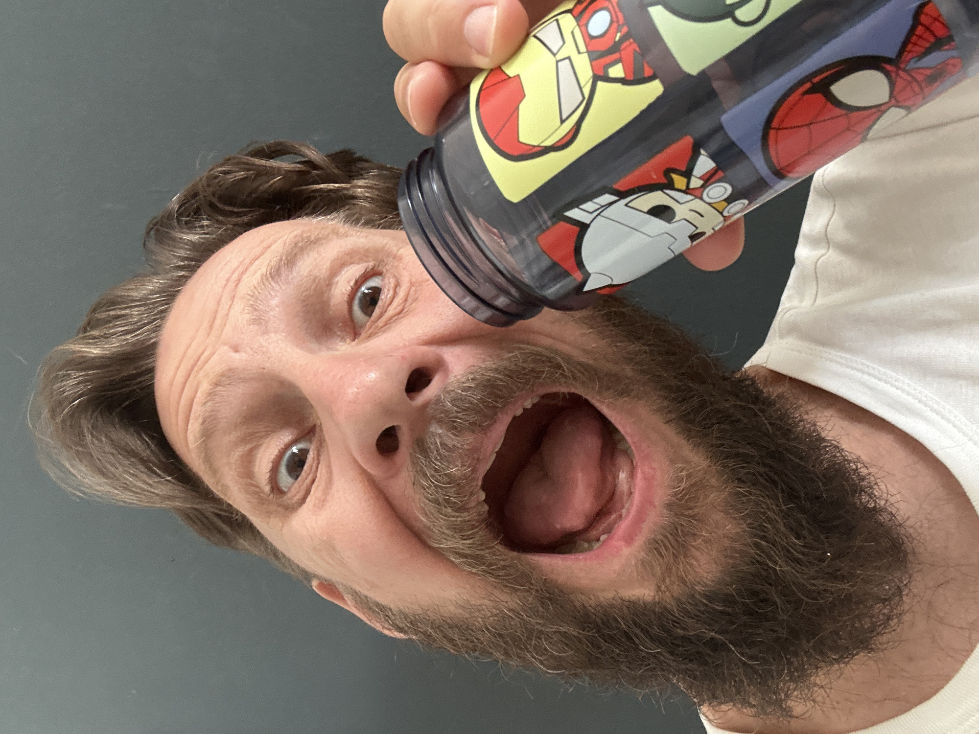 The image is a close-up, low-angle shot, rotated counter-clockwise, featuring a man in a playful and exaggerated pose. He looks directly at the camera with wide, dark brown eyes and an open mouth, revealing his tongue and the inside of his mouth. He has light brown, wavy hair and a prominent dark brown beard. Wrinkles are visible on his forehead, indicating an expressive facial movement. He wears a plain white t-shirt, parts of which are visible on the right side of the frame.
The man holds a clear plastic drinking bottle featuring colorful cartoon illustrations of popular superheroes, including Iron Man, Thor, and Spider-Man. The bottle is held upside down over his face, with its open end positioned near his left eye and forehead, suggesting a humorous attempt to drink or pour its contents.
The background is a plain, dark grey wall, indicating an indoor setting. The overall mood is lighthearted and comical, conveyed by the man's exaggerated expression. There is no discernible text on the bottle, only the stylized superhero characters. The location context provided for this image is Fethiye, Turkey.