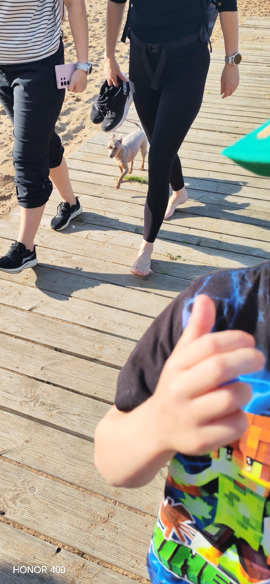 The image shows an outdoor scene on a sunny day, likely in the afternoon, judging by the long shadows cast. The ground is a wooden boardwalk, with sand visible on the left side, suggesting a beach or a waterfront area. In the foreground, a child's arm and shoulder are visible, wearing a colorful t-shirt with a pattern that resembles characters from the video game Minecraft. The child's hand is in the frame, giving a thumbs-up gesture.
Further back on the boardwalk, two people are walking. The person on the left is wearing black pants and a striped t-shirt. They are holding a pink smartphone in their left hand and have a white smartwatch on their right wrist. They are wearing black sneakers. The person on the right is wearing black leggings and a black top, also with a smartwatch on their right wrist. They are carrying a pair of black Hoka sneakers in their right hand. Between the two people, a small, light-colored dog with short fur is walking. The scene appears to be relaxed and casual, perhaps a stroll along the beach. The text "HONOR 400" is visible in the bottom left corner of the image, which is likely a camera or phone model designation.