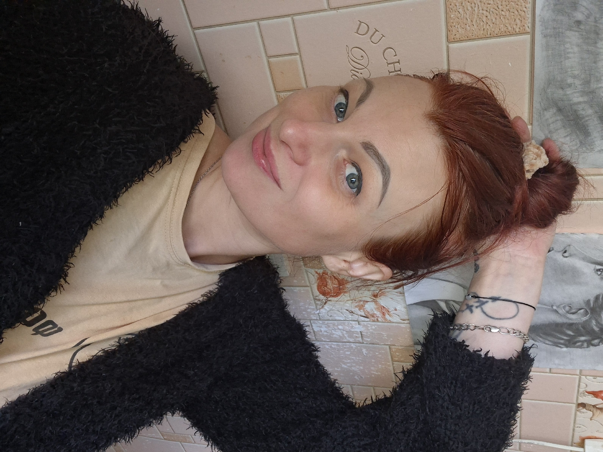A woman with auburn hair pulled into a bun is shown from the chest up. She is looking directly at the camera and smiling faintly. She has blue eyes and is wearing a black fuzzy cardigan over a tan t-shirt. There are tattoos on her left arm and a silver bracelet. The background appears to be a wall with pink tiles. There are also some artistic drawings or prints on the wall behind her. The scene suggests an indoor setting, possibly a home. There is no visual information available to indicate the specific location within Myadzyel, Belarus.
