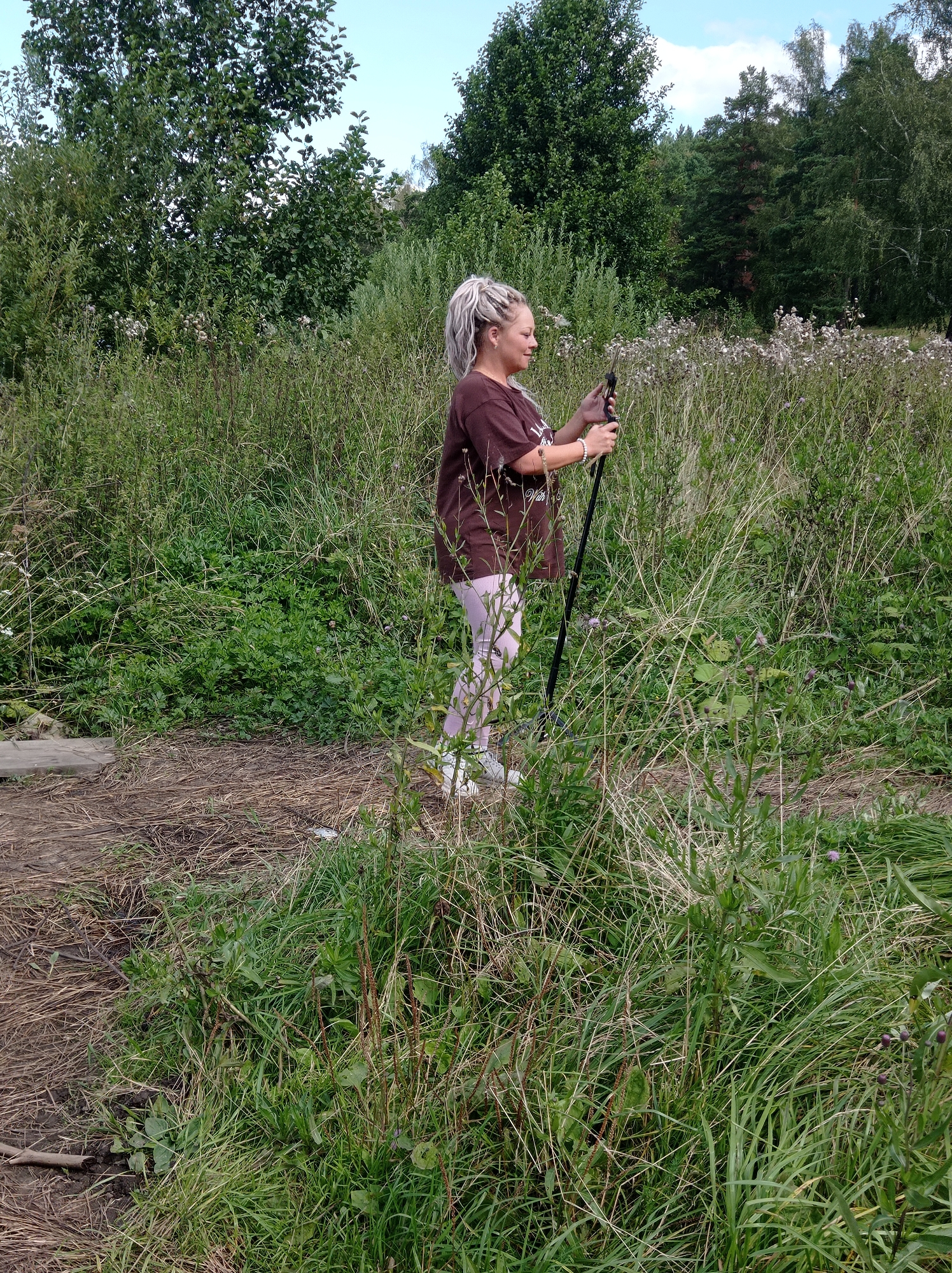 A woman with blonde dreadlocks stands in a grassy field holding two hiking poles. She is wearing a brown t-shirt and light pink leggings. The field is overgrown with tall grass and some scattered wildflowers. In the background, there is a dense line of trees and a clear blue sky with a few clouds. The ground in the foreground is covered with dry grass and some dirt. The location is the city of Degtyarsk, Russia.