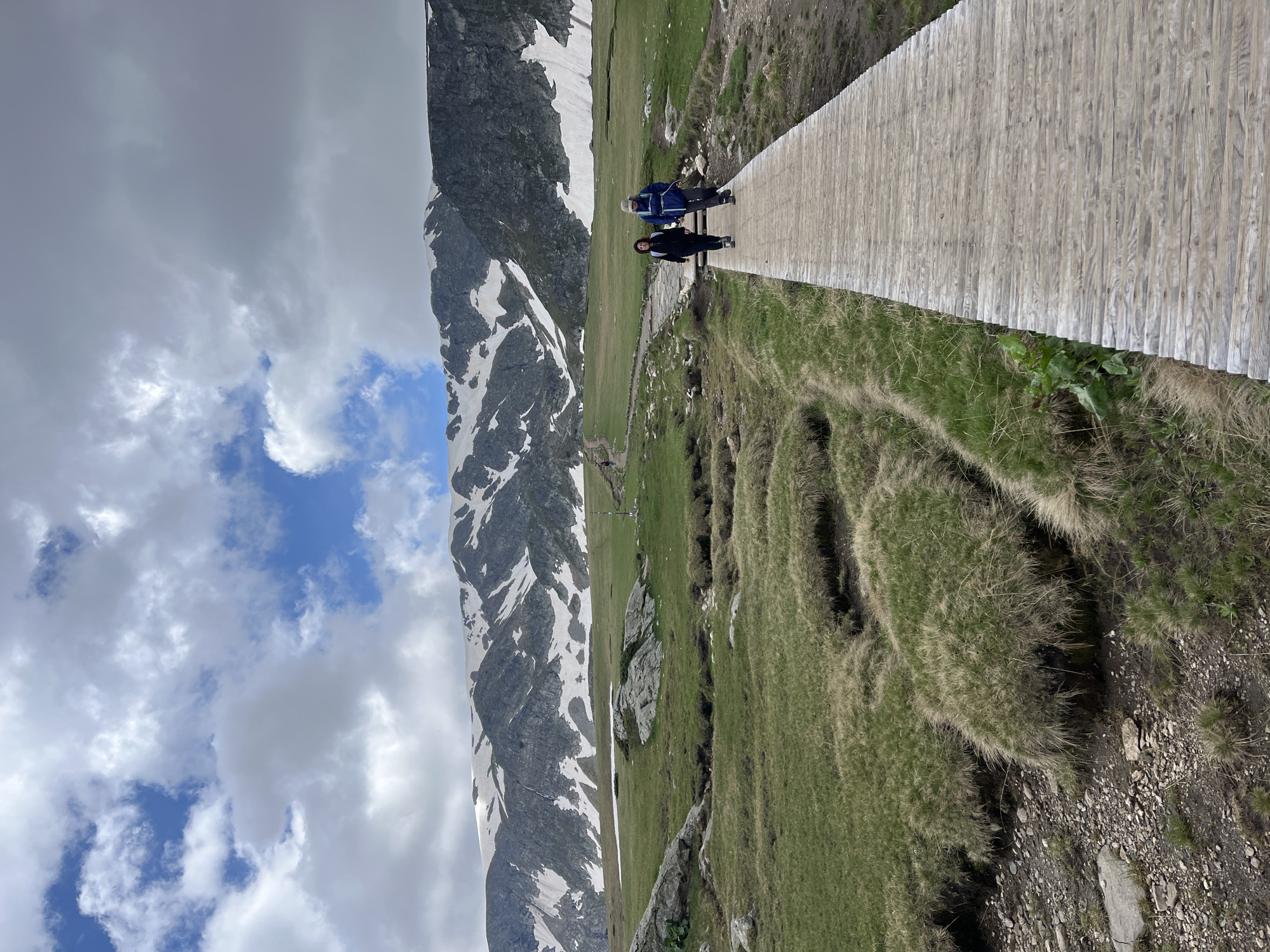 Two individuals are walking on a wooden boardwalk in a mountainous, grassy landscape. The person on the left is wearing a dark jacket and dark pants, and the person on the right is wearing a blue jacket and dark pants, with a light-colored hat. They are walking away from the viewer on the boardwalk, which runs from the lower right corner towards the center left of the image. The environment features green grass, some rocky areas, and a stream or ditch to the left of the boardwalk. In the background, a large mountain range is visible, with snow patches on its slopes and rocky outcrops. The sky above is partly cloudy with patches of blue sky. The scene appears to be in an outdoor, natural setting, likely a hiking trail.