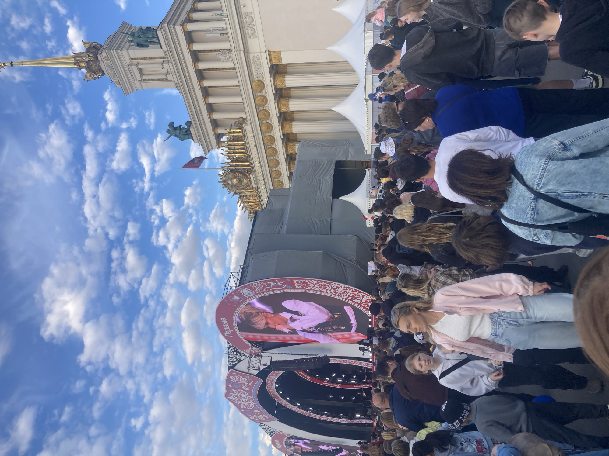 This image captures an outdoor public event, rotated 90 degrees to the left. A large crowd of people of various ages is gathered, facing a prominent stage area with large video screens. Many individuals in the foreground and midground are looking towards the stage, suggesting they are watching a performance or presentation.
In the background, a majestic cream-colored architectural building stands under a bright blue sky dotted with scattered white clouds. The building features classical columns, ornate golden decorations, and a distinctive golden spire, identifiable as the Central Pavilion (Pavilion No. 1) at VDNKh in Moscow, Russia. A red flag with an emblem flies from a pole near the building.
The stage setup includes a large circular video screen prominently displaying a female singer or presenter holding a microphone. Other stage structures are partially covered by gray tarpaulins, and white tent structures are visible behind the crowd, suggesting event infrastructure. The bright daylight indicates it is daytime with good weather.
The Cyrillic word "Первые" (Pervye), meaning "The First" or "First Ones," is clearly visible on the large circular screen and other decorative elements of the stage. The event appears to be a public gathering, possibly a concert or festival, attracting a significant number of attendees.