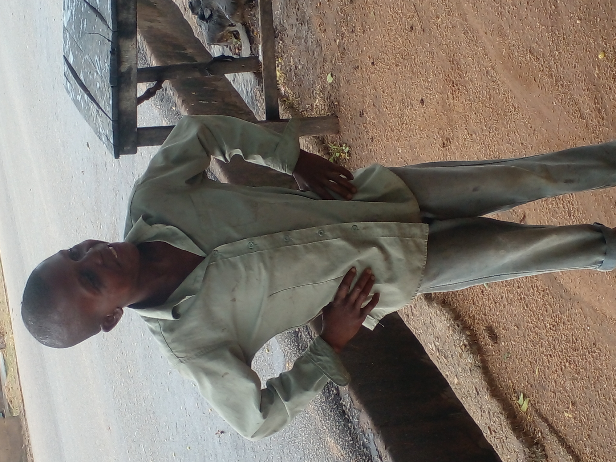 A young person with a shaved head stands outdoors, looking to their right with a smile. They wear a pale green collared shirt with the sleeves rolled up and grey pants. Both hands are placed on their hips. The ground is sandy and covered with small, light-colored particles. In the background, a weathered wooden table stands on legs, and a dark, textured surface runs along the lower left of the frame. The person appears to be in a casual setting, possibly on a street or a dusty area.