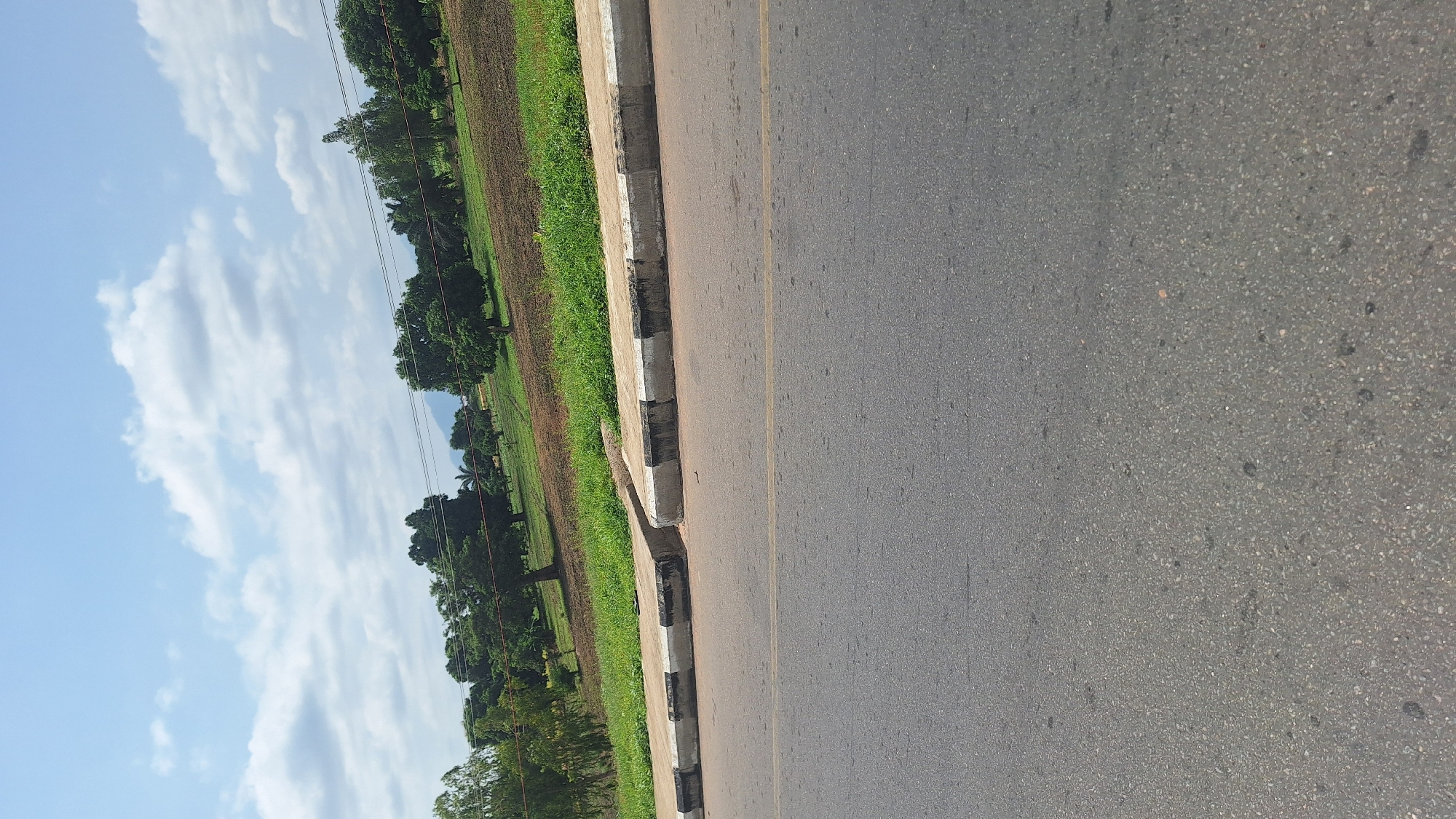 The image depicts a roadside scene in Jalingo, Nigeria, on a bright, cloudy day. In the foreground, a grey asphalt road with a faint yellow line down the center occupies the lower half of the frame. Adjacent to the road is a concrete curb with alternating black and white stripes, showing signs of wear and tear. Beyond the curb, a strip of green grass lines the edge of a gently sloping field. This field is characterized by patches of brown soil, suggesting cultivation, and scattered green vegetation. Further back, a line of lush green trees forms a natural barrier. In the distance, a hazy outline of mountains can be seen against the sky, which is filled with scattered white clouds. The overall impression is of a rural or semi-rural area on the outskirts of the city. No people or specific actions are visible in the frame.