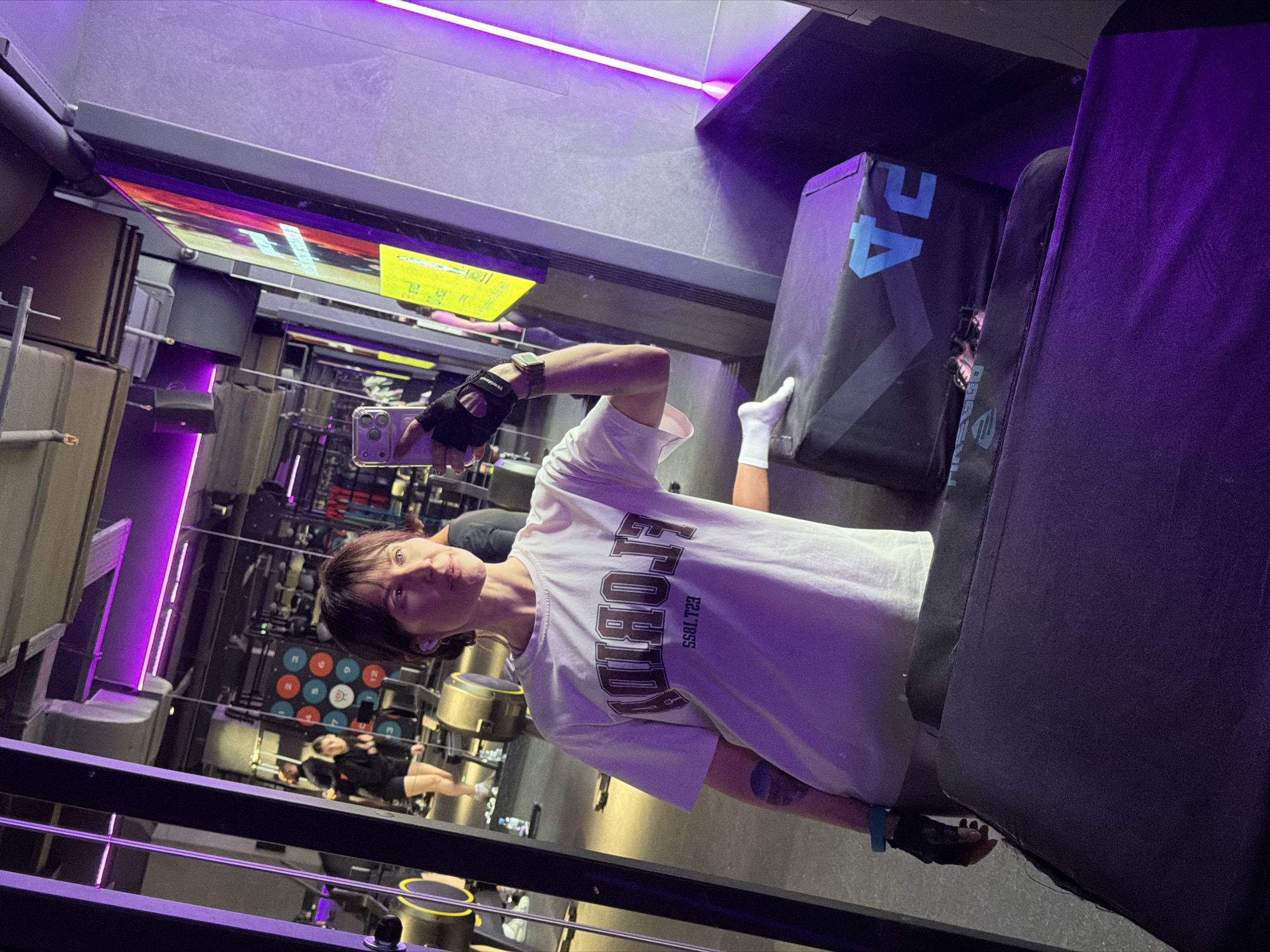 The image displays a person taking a selfie inside a modern gym or fitness studio in Perm, Russia. The individual has short, dark hair and a neutral expression, holding an iPhone with multiple cameras to capture the moment. They are wearing a white t-shirt with the inverted text "FLORIDA EST. 1853" visible on the chest, black fingerless workout gloves, and white ankle socks. A fitness tracker watch is worn on the left wrist, and a blue wristband is on the right.
The gym environment is illuminated predominantly by striking purple LED strip lighting along the ceiling and walls, creating a distinctive ambiance. In the background, various pieces of gym equipment are visible, including punching bags or mats marked with "2A" in light blue text, a weights rack with colorful circular weight indicators, and exercise steps or boxes. Reflections in the mirrored walls offer glimpses of other parts of the gym and another person, possibly working out, in the distance. A blurry reflection of a colorful screen or sign with yellow text is also visible. The scene suggests an indoor setting, likely during an evening or nighttime workout, given the artificial lighting.