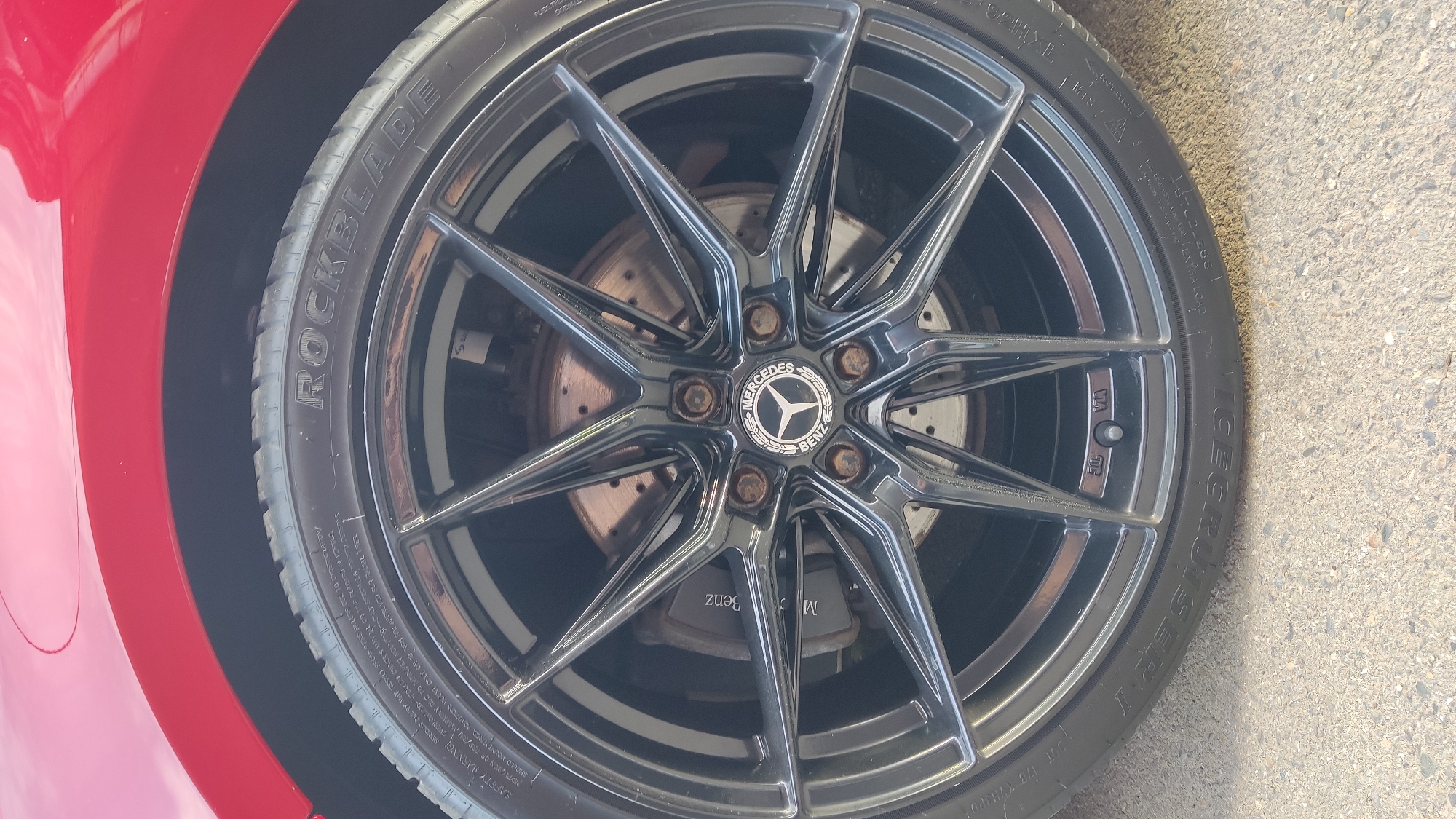 A close-up, eye-level shot captures the front wheel of a red Mercedes-Benz vehicle, likely parked outdoors on a sunny day in Krasnodar, Russia. The car's vibrant red fender is visible on the left side of the frame, curving around the wheel. The wheel itself is a prominent black multi-spoke alloy rim with a high-gloss finish, reflecting the surroundings.
At the center of the wheel, a silver Mercedes-Benz logo is clearly visible against a black background, encircled by the words "MERCEDES BENZ". The five lug nuts around the center cap show signs of rust. Behind the spokes, a perforated and vented brake rotor is discernible, along with a brake caliper which also appears to have "Mercedes Benz" text on it, albeit inverted in the reflection.
The tire is a "ROCKBLADE" brand, with "ICECRUISER I" also embossed on its sidewall, suggesting it might be a winter tire. Other legible text on the tire includes "SAFETY WARNING", "ROTATION", and "EXTRA LOAD". The tire tread is visible, and the tire appears to be in good condition. The vehicle is positioned on a light gray, textured concrete or asphalt surface, which occupies the right and bottom portions of the image. The bright sunlight creates clear reflections on the glossy wheel and tire. No people or specific activities are depicted, focusing solely on the vehicle's wheel assembly.