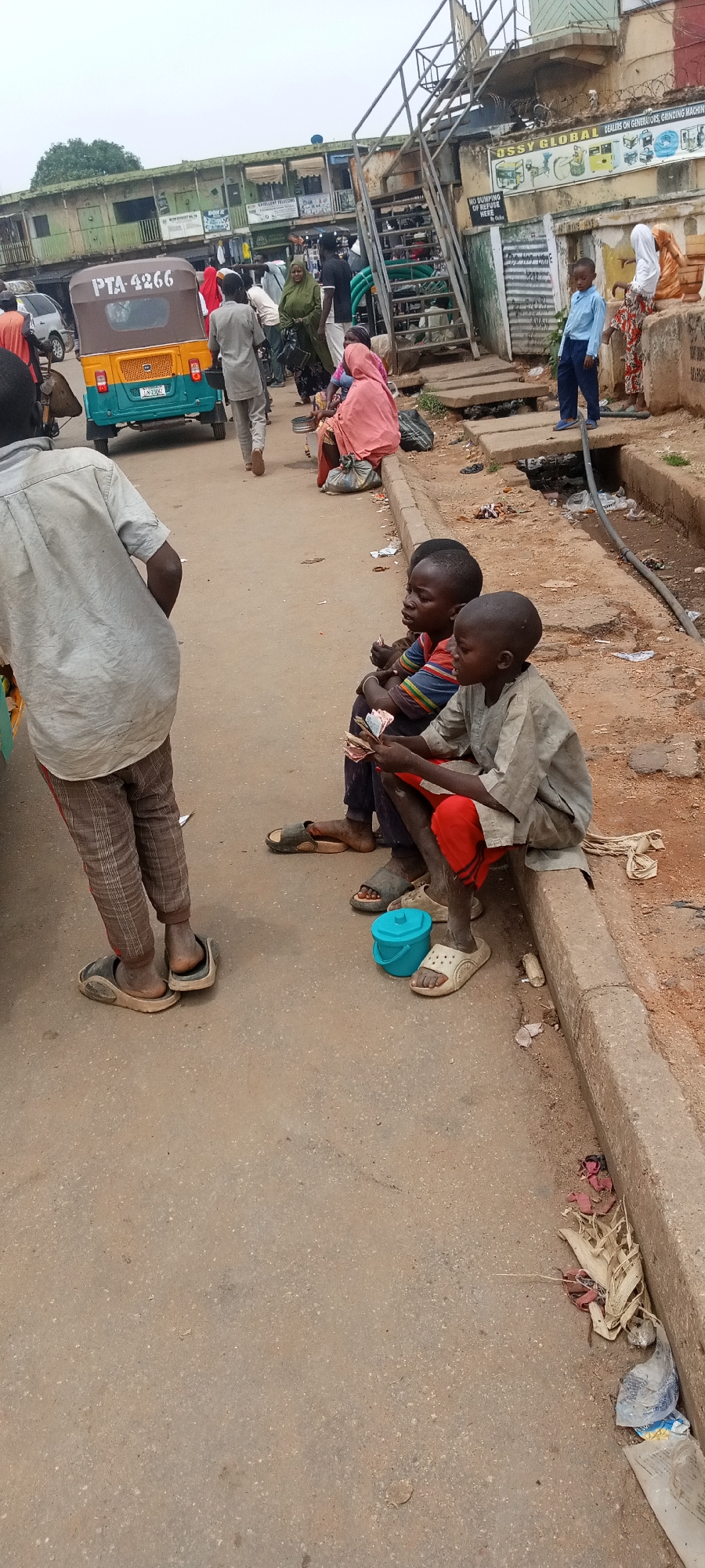 The image shows a street scene in Jos, Nigeria. Two young boys are sitting on a curb next to the road, holding money. There is a small, light blue plastic bucket next to them. A person in a grey shirt and patterned pants stands nearby, with their back to the camera. Other people are walking on the street and sitting near a covered drainage ditch. A green and yellow tricycle taxi with the license plate PTA-4266 is visible in the background. There are buildings lining the street, some with signs, and an elevated pedestrian walkway with stairs. The environment appears to be urban and somewhat impoverished.