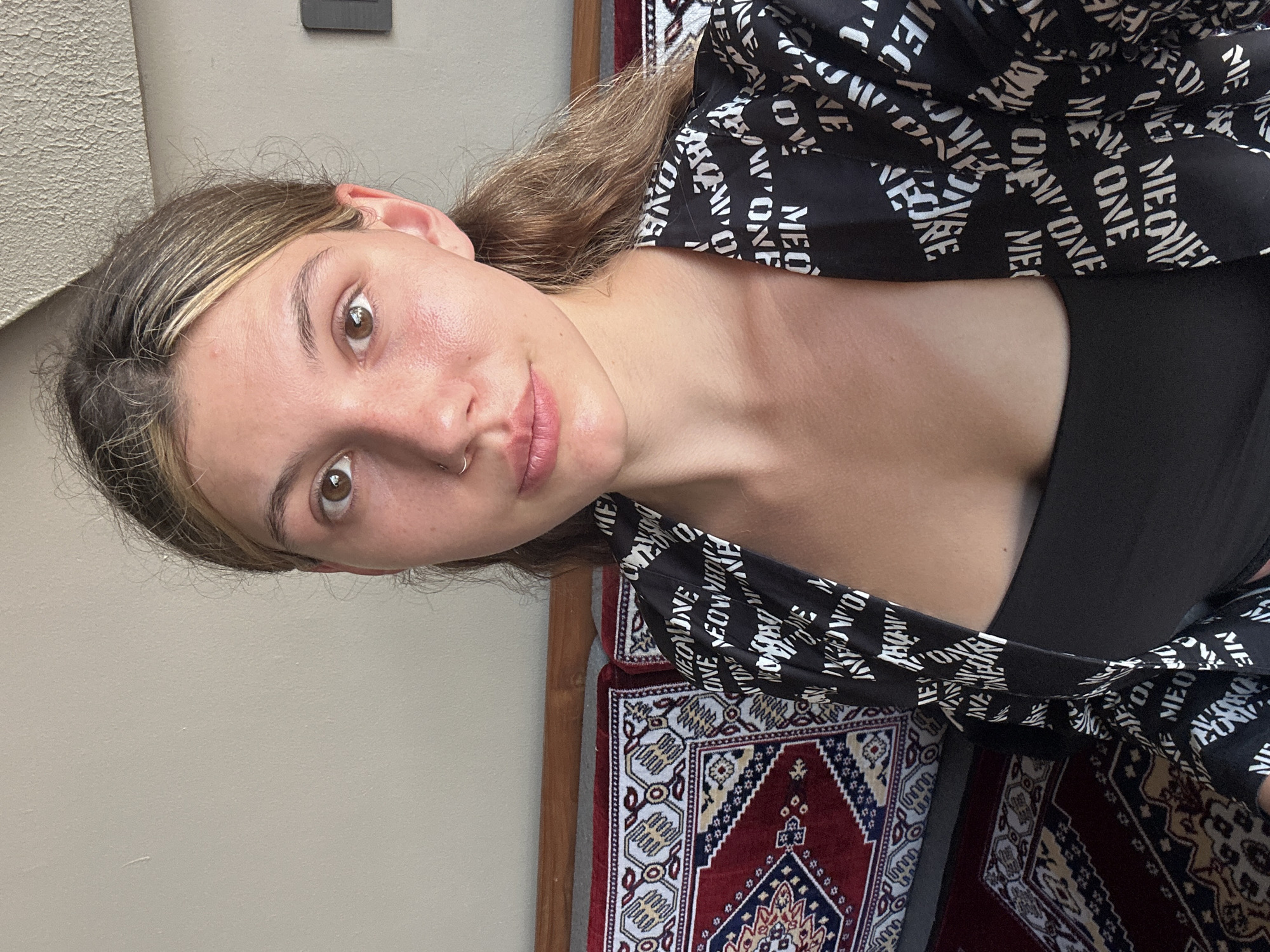 A young woman with brown hair and a nose ring poses for a selfie. She wears a black top and a black and white patterned shirt with text "MEOWMEONE". She is indoors with a wall on one side and a patterned rug on the other. The location is unknown, and there is no indication of Baan Chaweng, Thailand.