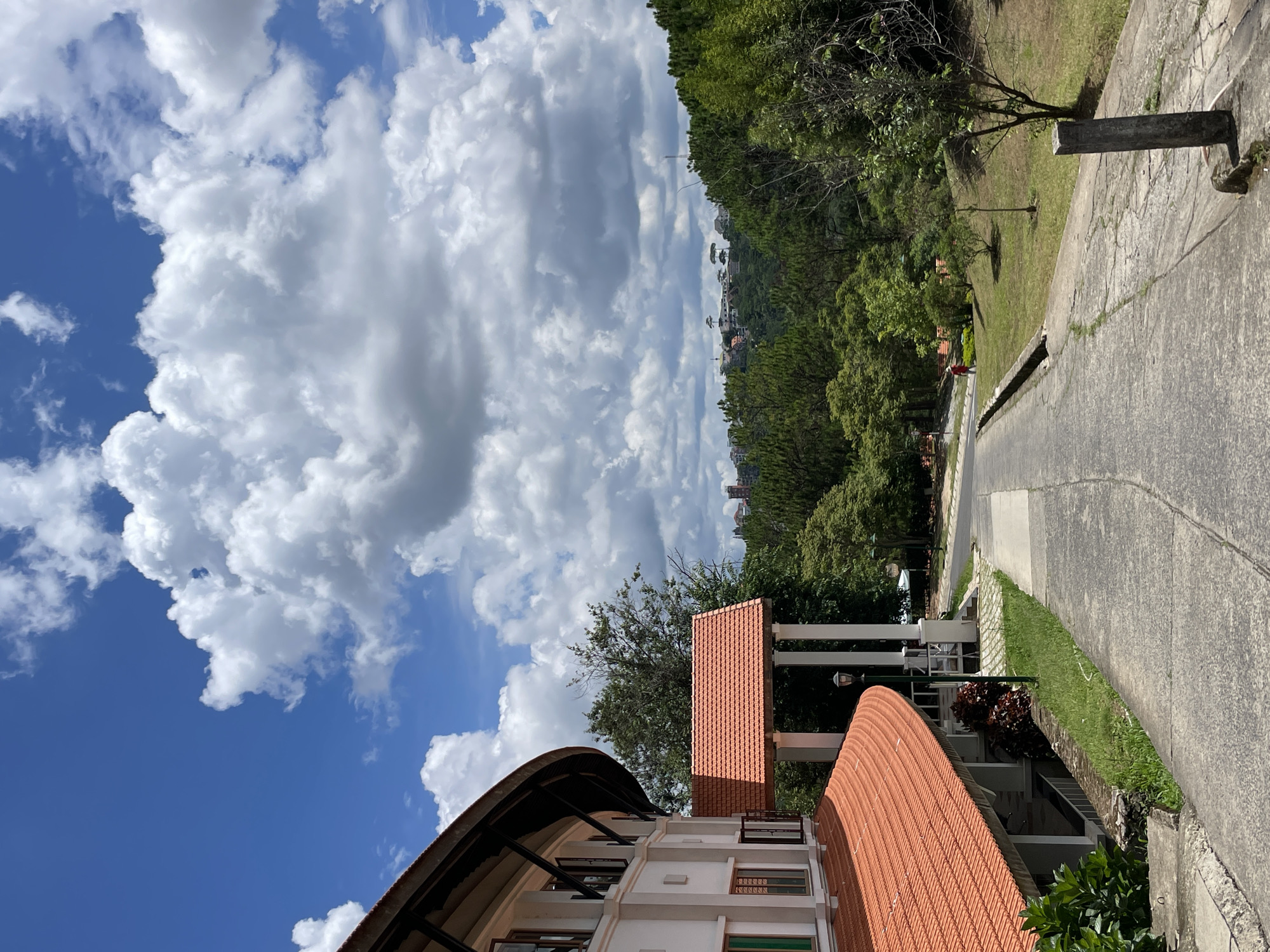 A paved pathway curves upwards through a grassy area with trees and a building on the left, leading towards a distant town nestled in a lush, green valley under a bright blue sky filled with dramatic white clouds. The building on the left features a curved, white facade with multiple windows and a striking orange tiled roof. A covered walkway with a similar orange roof is supported by white pillars. Further up the path, a lone individual in a red shirt walks away from the camera. The landscape is characterized by rolling hills covered in dense trees, with a cluster of buildings visible on a hilltop in the distance. The overall scene depicts a serene, sunny day in a hilly, possibly university or resort-like setting, likely Da Lat, Vietnam, given the distinctive architecture and landscape.