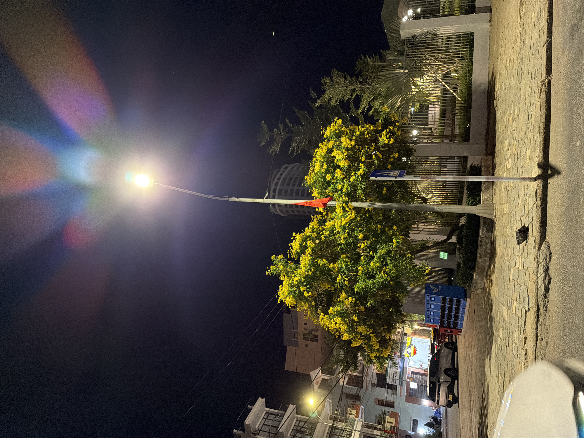 The image depicts a nighttime street scene in Nha Trang, Vietnam, illuminated by a streetlamp that casts a bright glow and creates lens flare with colorful light refractions. The foreground features a cobblestone sidewalk with patches of rough asphalt, indicating wear and tear. To the left, a silver SUV is parked on the street. A blue electrical charging station with multiple outlets is positioned on the sidewalk near a lush, vibrant tree with abundant yellow flowers. Behind the tree, a metal fence encloses a landscaped area with palm trees and decorative lighting. Several multi-story buildings with visible windows and balconies line the street on the left, with one building exhibiting a curved architectural design in the background. A red flag is affixed to a pole near the yellow-flowering tree. The night sky is dark, with a few faint light sources visible.