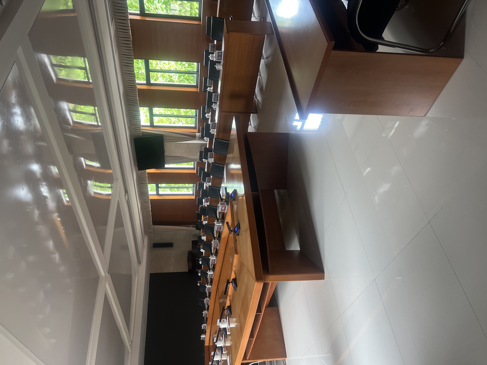 The image shows the interior of a conference room in Jiangjin, China. The room is dominated by long, wooden tables arranged in rows with chairs placed at each setting. Each setting is equipped with a microphone. Large windows are visible along one side, providing natural light and a view of greenery outside. A screen is visible at the end of the room. The room appears unoccupied and ready for a meeting or conference.
