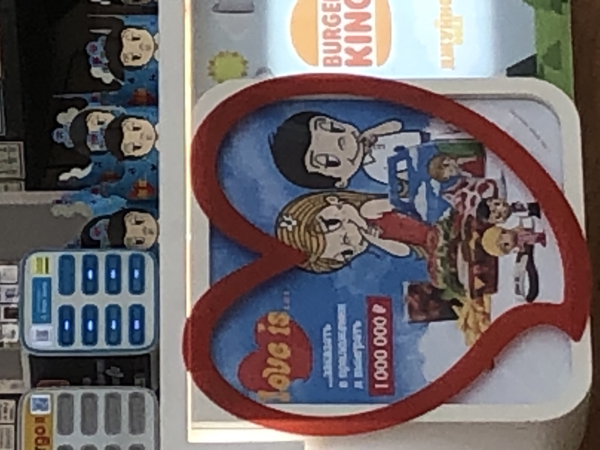 The image displays a promotional setup within a Burger King fast-food restaurant in Krasnoyarsk, Russia.
In the foreground, a white rectangular display board is framed by a red, textured, heart-shaped border. The board features an illustration on a blue background with white clouds. On the upper left, the text "Love is..." is rendered in stylized orange letters. Below it, in Cyrillic, the text reads "ЗАКАЗАТЬ В ПРИЛОЖЕНИИ И ВЫИГРАТЬ 1 000 000 Р" (ORDER IN THE APP AND WIN 1,000,000 ₽). The illustration depicts a blonde, cartoon-style woman in a red top on the left, and a dark-haired, cartoon-style man in a white polo shirt on the right, both with hands near their chins. Between them, a table is laden with fast food, including a large hamburger, french fries, and a dark beverage. Below the table, two small figurines—one blonde in pink, one dark-haired in white—are visible, alongside a small grey object resembling a gaming controller or toy guitar.
To the right of this heart-shaped display, a partial "BURGER KING" logo is visible, with the word "BURGER" on an orange semi-circle and "KING" below it. Faint Cyrillic text is discernible beneath the logo, likely a product name.
In the upper portion of the image, behind and above the heart display:
On the far left, a light-colored vertical charging station with multiple grey rectangular slots is partially visible.
Adjacent to it, a blue and white charging station or locker unit features two columns of four rectangular slots each, some illuminated with blue lights. A QR code or similar icon is present on its top left.
To the right of this unit, several stylized blue and white cutouts, resembling abstract castles or clouds, are visible. Four cartoon faces, similar in style to the "Love is..." characters, peek over these cutouts. These faces have varying hair colors and styles.