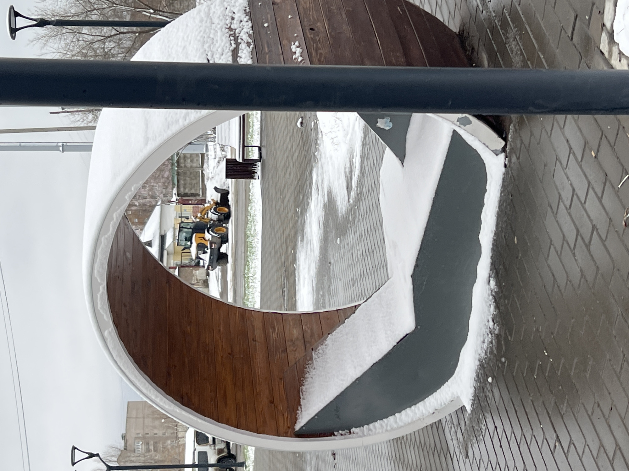 The image shows a circular bench structure in Chaikovsky, Russia, partially covered in snow. The bench is made of wood and gray metal. It is located on a brick-paved surface. Through the bench's opening, a construction vehicle can be seen in the distance. A black metal pole is in the foreground, slightly obscuring the view. Street lamps are also visible in the background.