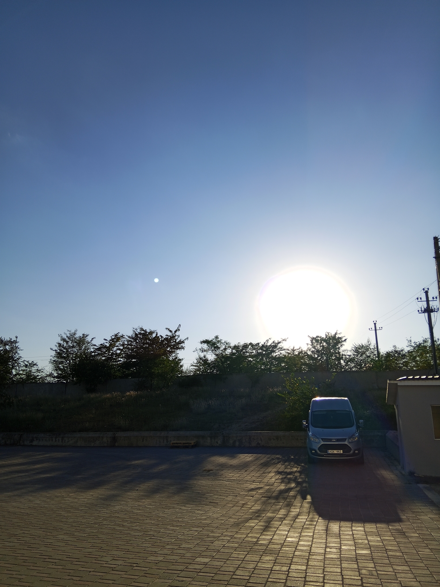 The image depicts an outdoor scene bathed in the bright, low light of the sun. The sky is a clear, gradient blue, transitioning to a lighter hue towards the horizon. A prominent, overexposed sun appears in the upper right quadrant of the sky, with a bright halo. To the left of the sun, a smaller, more defined bright spot is visible, possibly the moon or a lens flare.
The foreground is a paved area with interlocking brickwork, casting long shadows from unseen objects. In the middle ground, a line of trees and shrubbery forms a natural barrier. Behind the trees, a concrete retaining wall is visible.
A silver-colored van is parked in the paved area, facing the viewer. The van's license plate reads "KQX 983". To the right of the van, a portion of a building with a light-colored wall and a dark-pitched roof is visible. Electrical poles with wires are also present in the scene, indicating a developed area. The overall impression is a quiet, possibly rural or suburban setting at either sunrise or sunset.