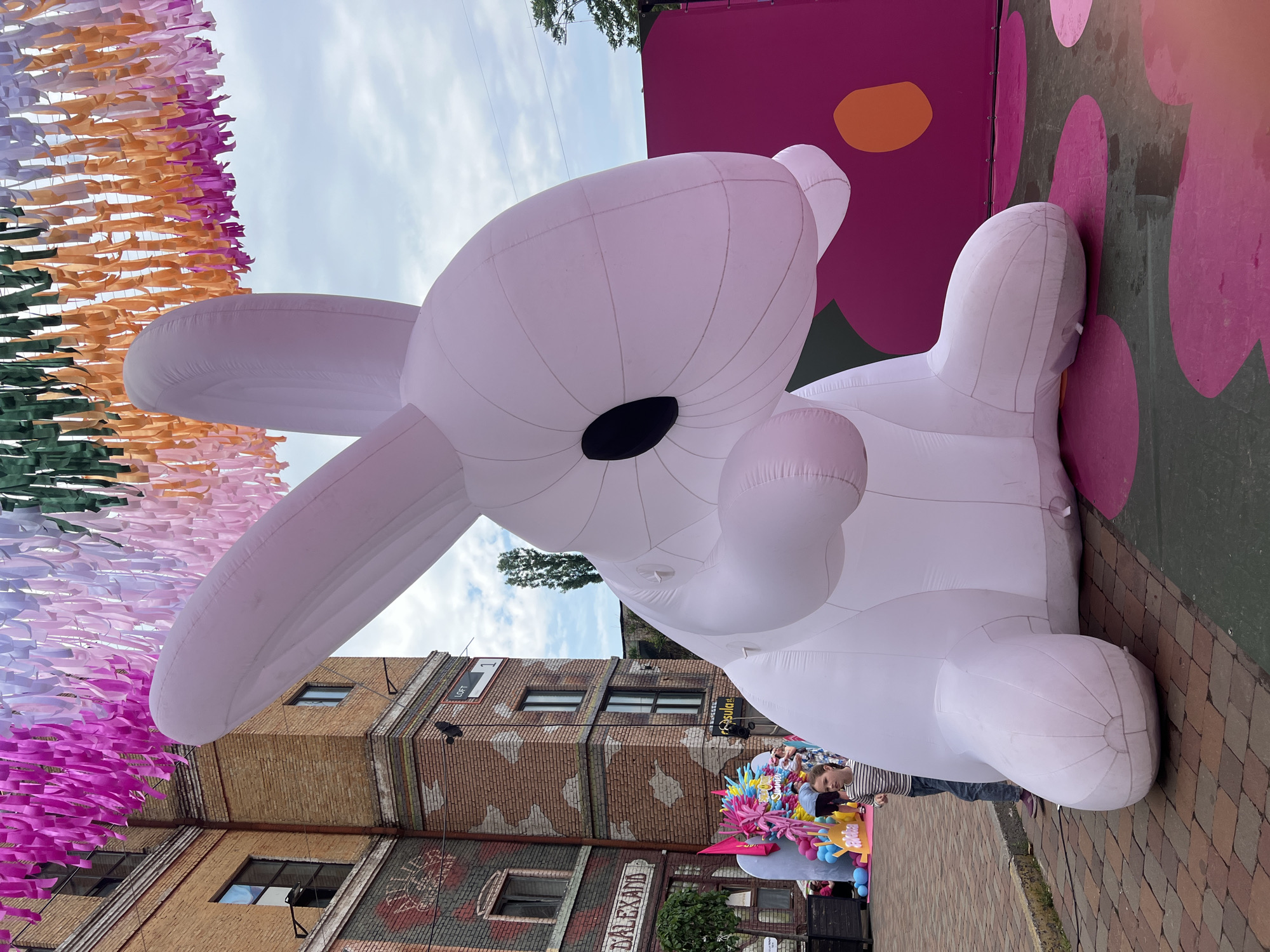 A large, white, inflatable rabbit sculpture, depicted in a seated position with prominent black eyes and rounded limbs, occupies the central and right portions of an outdoor scene. The rabbit faces right. To its left, a young girl, approximately 7-10 years old, wearing a striped long-sleeved shirt and patterned pants, stands observing the sculpture.
The foreground features reddish-brown brick paving under the girl and the rabbit's front paws, transitioning to a dark gray painted surface with large, irregular pink and orange circular motifs under the rabbit's rear.
In the background on the left, multi-story brick buildings with large windows are visible. The leftmost building features a partial mural or sign that includes "D.ALEXAND" and "ATELIER." Adjacent to it, a central brick building displays a white rectangular sign with "LOFT" above "1" in red text. Further back, on the far left, indistinct, colorful decorations and additional figures are visible, including a blue and pink sculpture and a sign partially reading "Akiba."
Overhead, numerous colorful fabric streamers or ribbons, ranging from vibrant pinks and purples to whites, greens, and oranges, are strung across the entire width of the image, creating a canopy. A partially cloudy sky is visible through the gaps in the streamers. On the right side of the frame, behind the rabbit, a solid magenta-pink wall or backdrop with an orange circular element is present. Overhead power lines and distant tree foliage are visible against the sky in the upper right.
The architecture, signs, and festive outdoor setting are consistent with the "Art Factory Platforma" (Арт-завод Платформа) creative cluster in Kyiv, Ukraine, known for hosting events and utilizing repurposed industrial buildings.