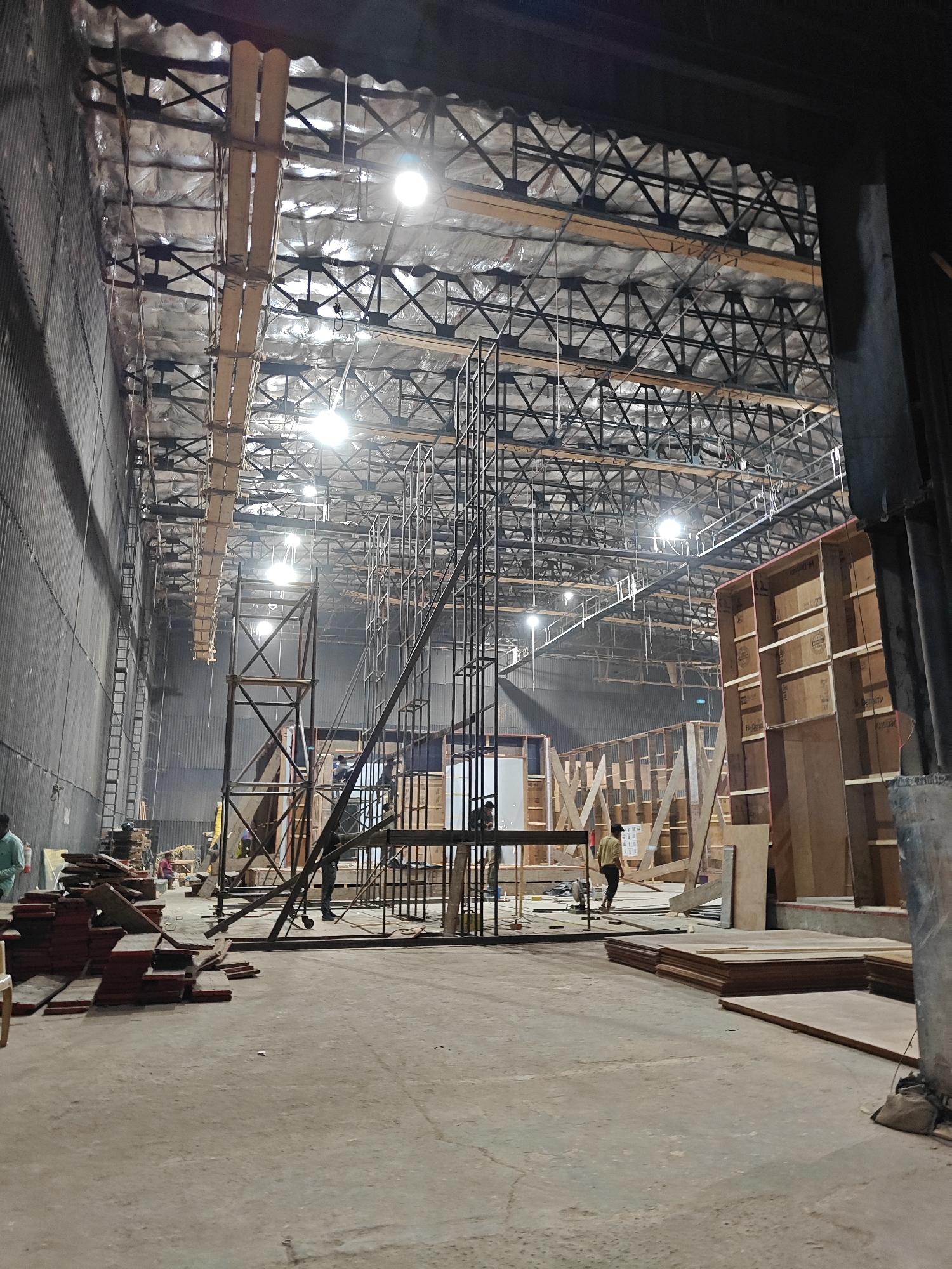 The image depicts a large film studio under construction in Mumbai, India. The expansive interior space features a high, exposed ceiling with a complex network of metal beams, insulation, and suspended lighting fixtures. Scaffolding structures of varying sizes and designs are scattered throughout the room, indicating ongoing set construction. Several individuals are present, some standing on scaffolding and others on the floor, possibly engaged in construction work. Construction materials, including stacks of plywood, wooden beams, and other building supplies, are visible on the concrete floor, contributing to the cluttered yet organized atmosphere of an active film set. The walls are lined with corrugated metal sheets, and sections of partially completed set walls are evident, suggesting a film set in the making.