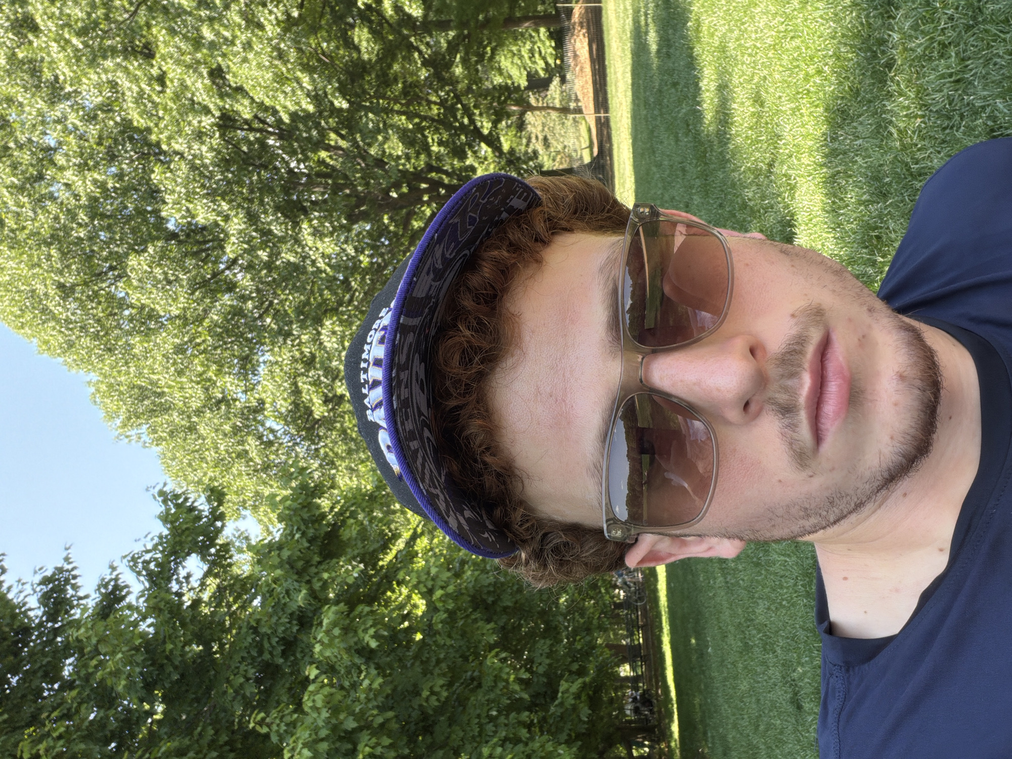A man lies on his back on a grassy lawn under trees, wearing sunglasses and a Baltimore Ravens baseball cap. The location is in New York, United States of America.
