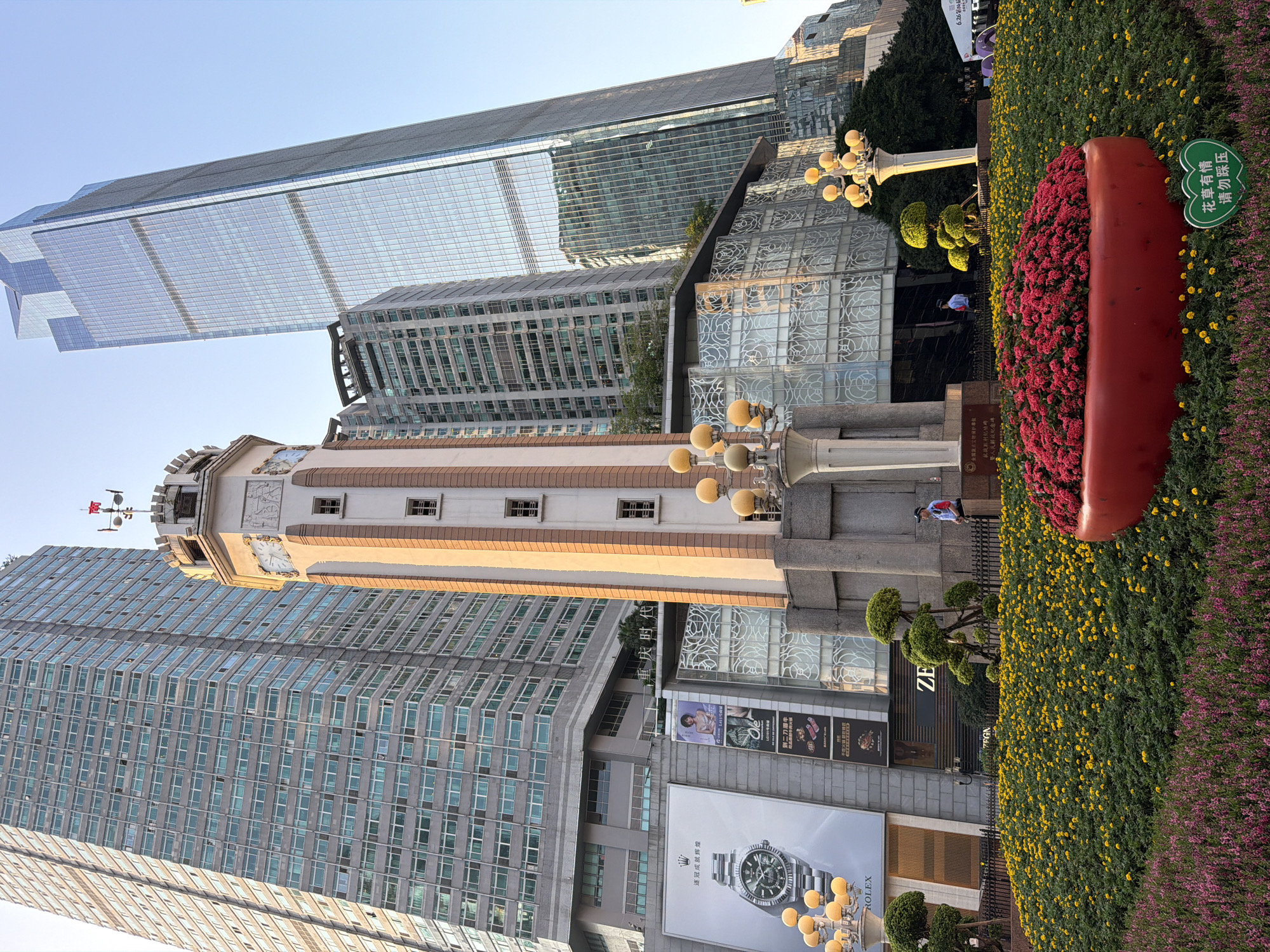 In Yuzhong District, China, a clock tower stands prominently in front of a backdrop of modern skyscrapers and older buildings. The clock tower, with its white and beige facade, features a visible clock face and decorative carvings. Surrounding the tower, a vibrant display of flowers, primarily yellow and pink, creates a lush foreground. A large, red planter filled with red flowers is a focal point within the floral arrangement. Two individuals are visible: one in a uniform standing near the clock tower, and another further back near a white tent. The scene is illuminated by natural light, suggesting it is daytime.