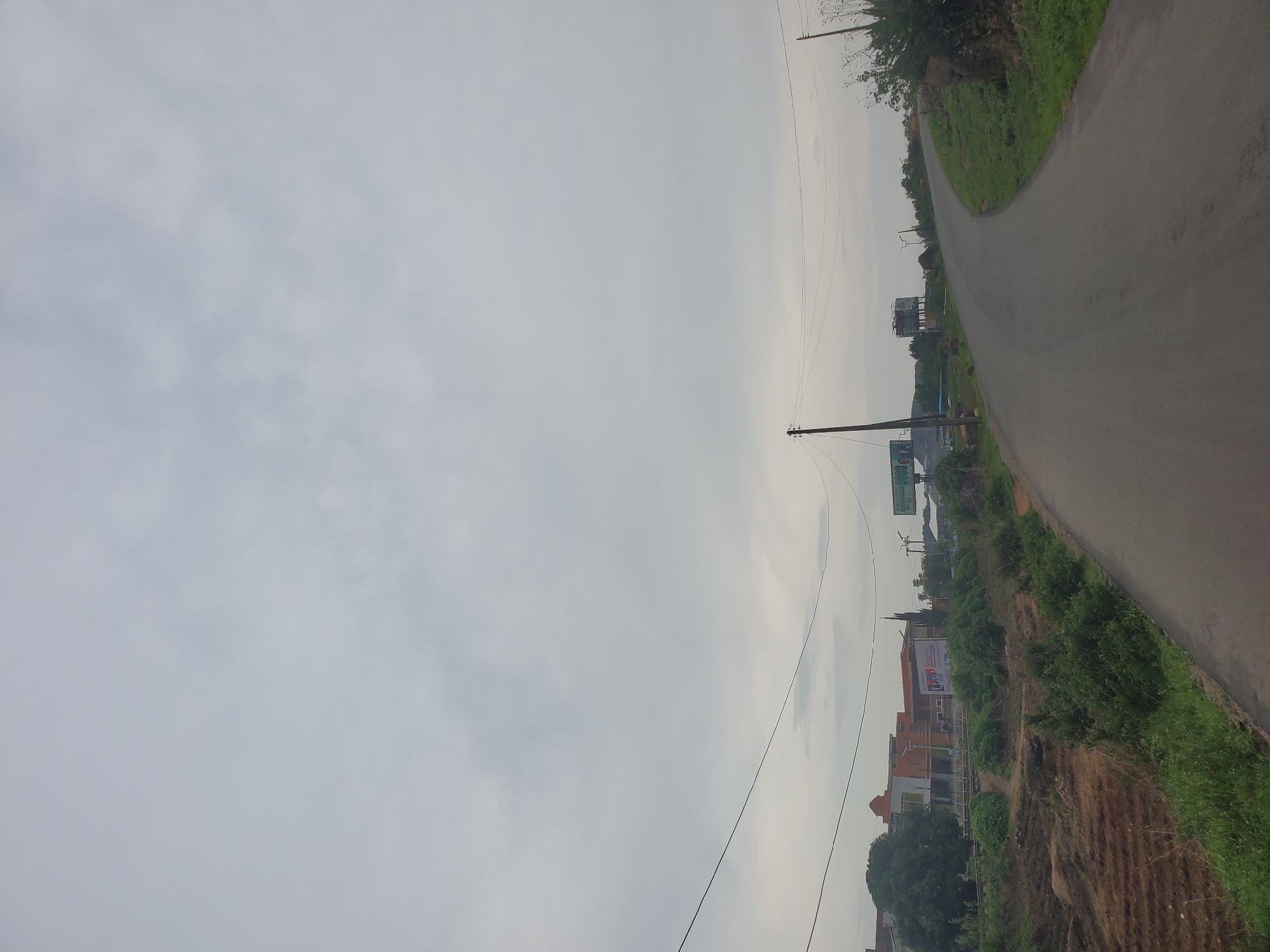 The image depicts a road curving through a slightly hilly landscape under an overcast sky. The road surface is asphalt, and it is flanked by grassy areas and some scattered rocks on the right side. On the left side of the road, there are cultivated fields with visible tilling patterns, followed by a line of vegetation and buildings. These buildings appear to be a mix of residential or commercial structures, with a large, multi-story building with orange accents and a billboard visible. Further down the road, there is a water tower and another billboard. Power lines stretch across the sky, supported by utility poles. The overall impression is of a semi-urban or rural setting, possibly on the outskirts of Jos, Nigeria, given the style of the buildings and the presence of utility infrastructure. There are no people visible in the image.