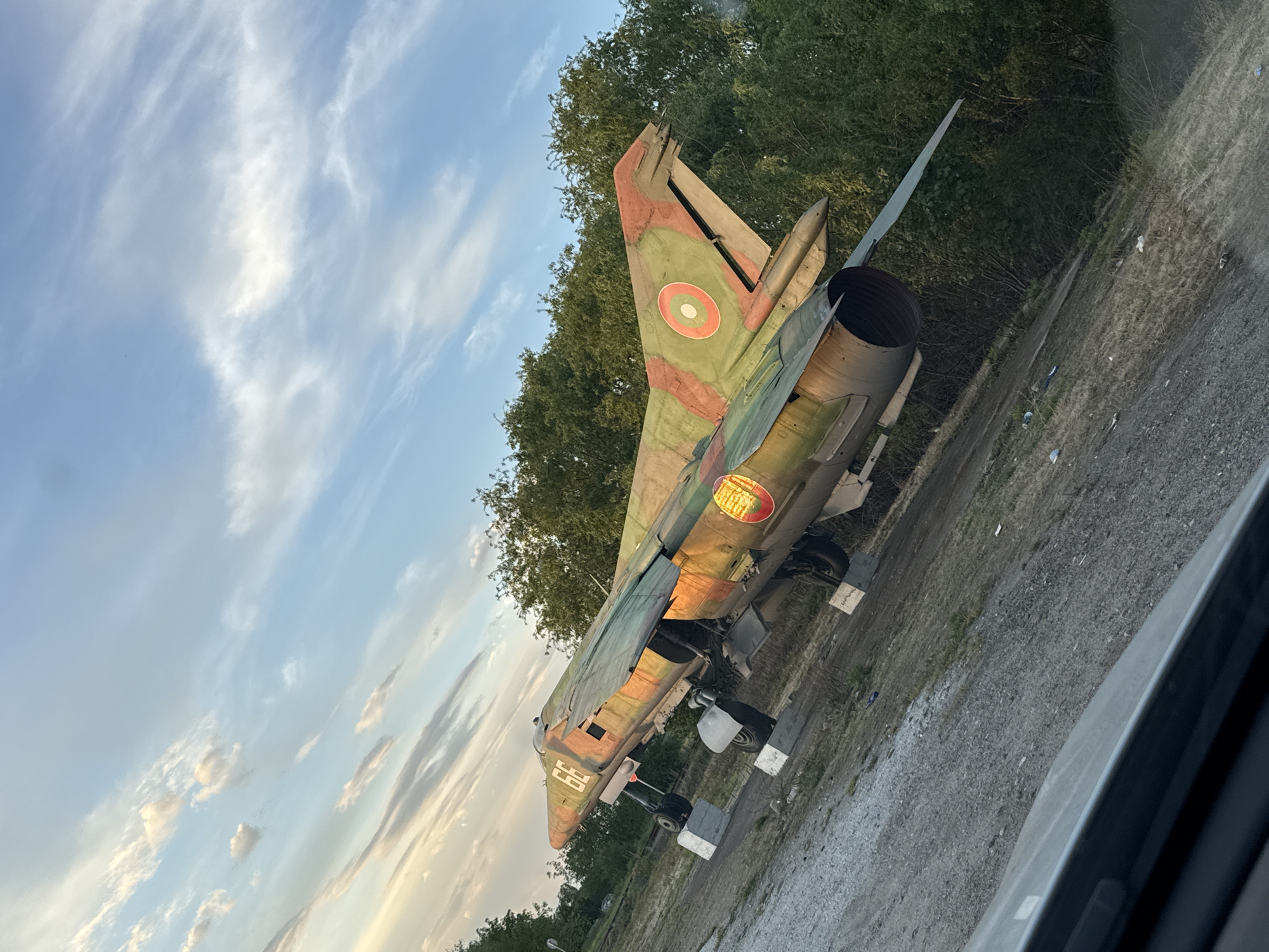 The image shows a disused MiG-23 fighter jet, likely a monument or display piece, situated in what appears to be a semi-rural environment. The aircraft is painted in camouflage colors and has the markings of the Bulgarian Air Force (red circle with white and green rings) on its tail. The jet is positioned on concrete blocks or low stands. The background consists of trees and sky with light clouds. The photo was taken in Cheshnegirovo, Bulgaria. The edge of a vehicle's interior is visible in the lower right corner, suggesting the photo was taken from a car.