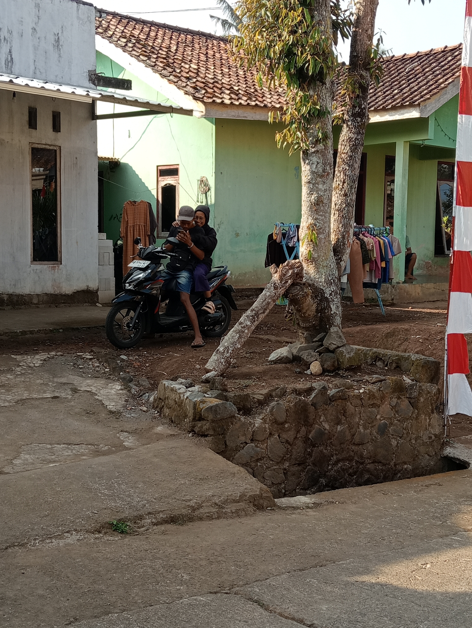 The image depicts a scene in what appears to be a village or suburban area, likely in Krajan Kidul, Indonesia, given the contextual information. In the foreground, a dirt and concrete pathway leads towards a paved area where a black motorcycle is parked. Two individuals, a man and a woman, are seated on the motorcycle. The man, wearing a cap and dark clothing, is looking down at a mobile phone. The woman, wearing a dark hijab and clothing, is seated behind him, also looking at the phone. They are both positioned on the rider's seat, with the woman's arms around the man.
Behind the motorcycle, a cluster of houses with terracotta tiled roofs and light green walls are visible. Clothes are hanging on a drying rack between two trees in the mid-ground. One tree has a bent trunk, suggesting it may be old or has experienced stress. The ground around the houses is uneven, with patches of dirt and stones. In the far right of the image, a partially visible Indonesian flag, with red and white stripes, stands tall. The overall lighting suggests it is daytime, possibly late afternoon, with shadows cast on the ground. The environment appears rustic and residential, with signs of daily life such as drying laundry and a common mode of transportation.