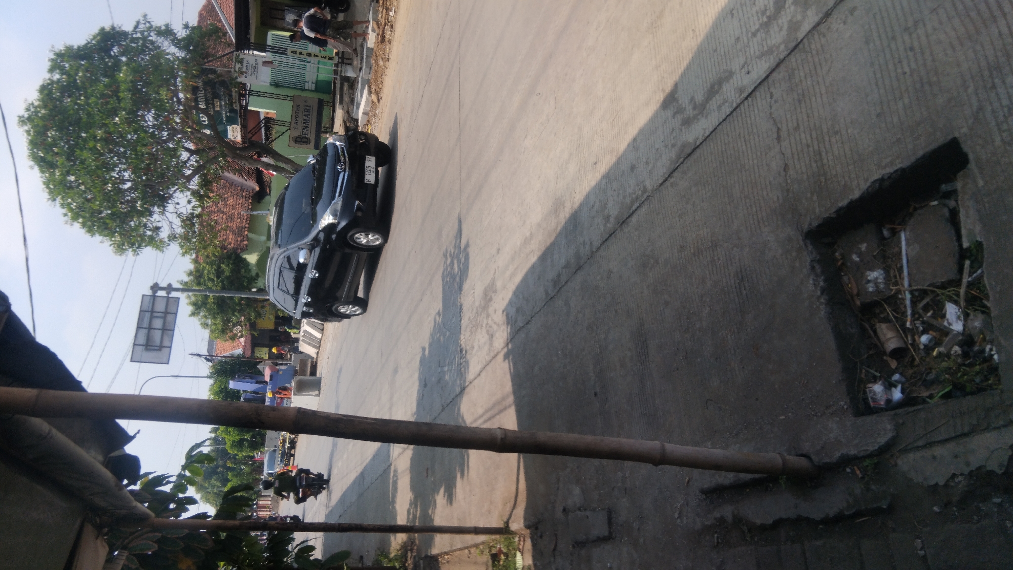The image shows a street scene in Batang, Indonesia, on a clear day. A black car is driving on the road, and a person on a motorcycle is visible in the background. The street has a sidewalk with a drainage grate that is filled with debris. There are buildings and trees visible on the side of the road. The overall impression is of a typical urban street in Indonesia.