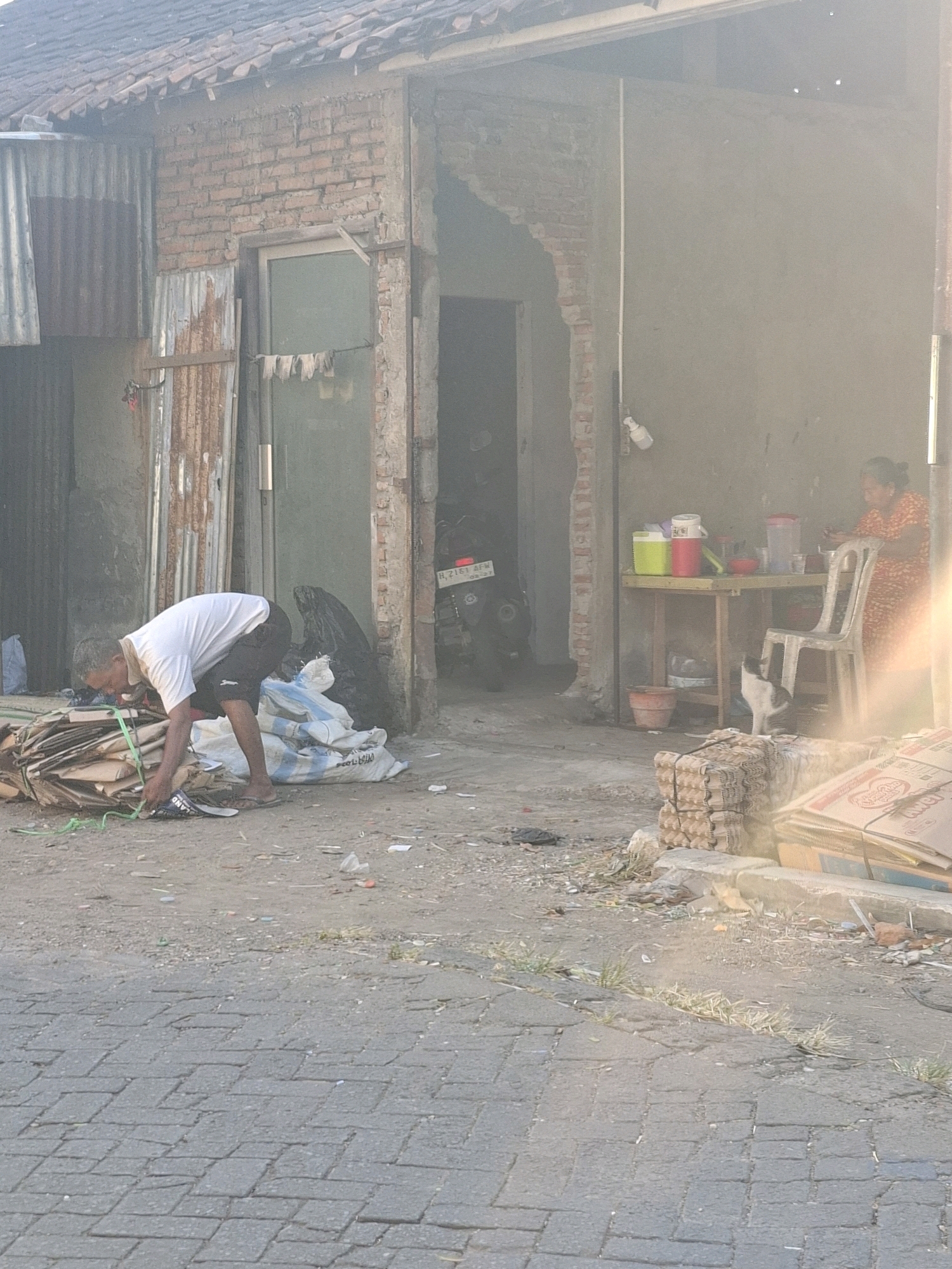 A man in a white t-shirt and black shorts is bent over, sorting through a pile of cardboard and plastic. He is wearing sandals and a light-colored face mask. To his right, there are several large sacks, one of which appears to be blue and white. Behind him, a motorcycle is parked. The man is in front of a brick building with a damaged section of wall. To the right of the building, there is an outdoor table with two chairs and various items on it, including containers and what appears to be food. A woman in an orange and white patterned dress is sitting at the table. A black and white cat is standing on the ground near the table. The scene appears to be in an urban or semi-urban environment, possibly a street or alleyway, with a paved surface in the foreground and debris scattered around. The overall impression is one of a working-class or informal economy setting. Given the context of Semarang, Indonesia, this could be a scene of waste recycling or a small roadside business.