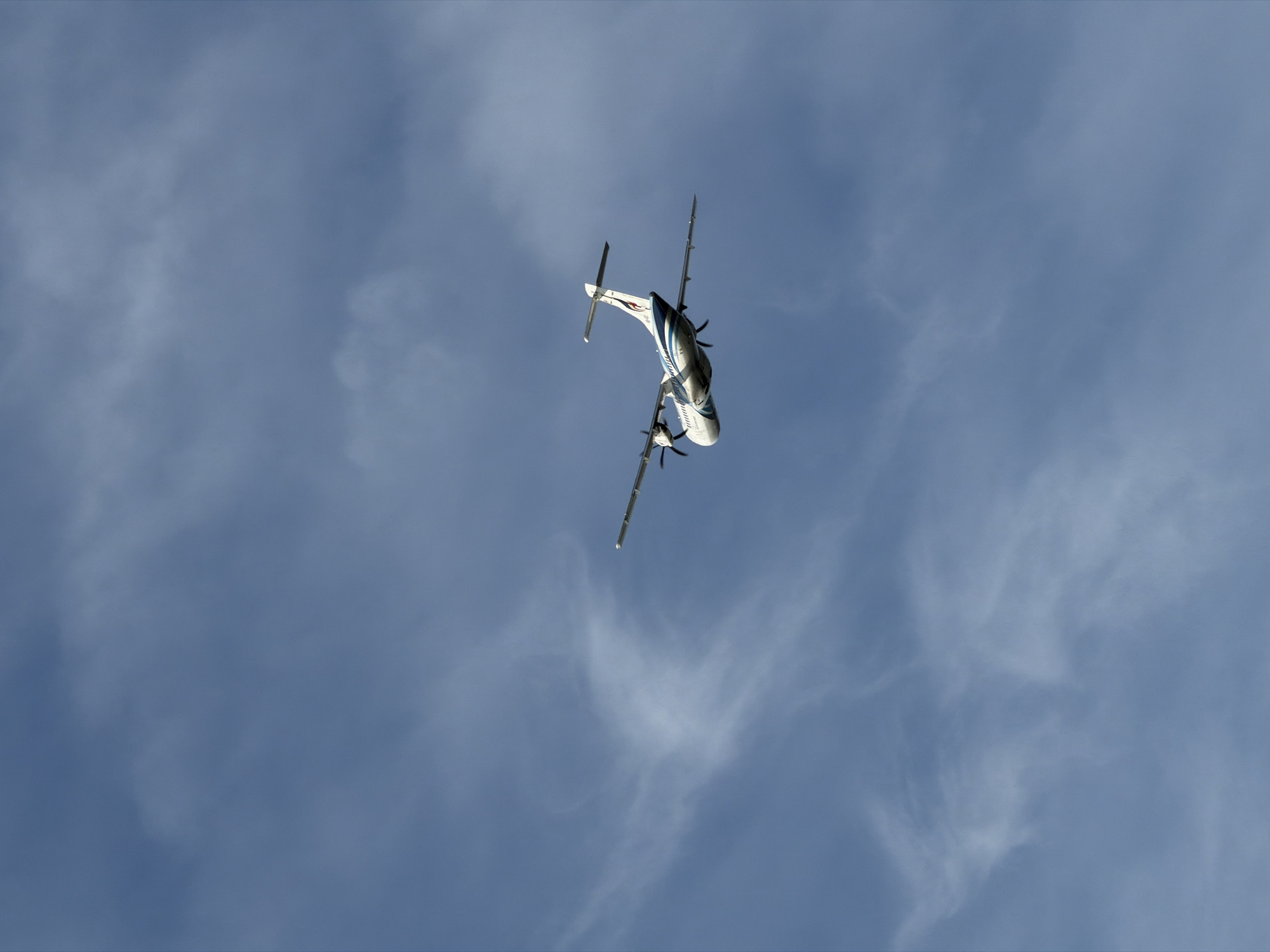 A low-angle, full shot captures a turboprop airplane against a backdrop of a blue sky filled with wispy white clouds. The airplane, predominantly white with blue accents, is seen from below and angled upwards, as if it is ascending or performing a maneuver. Its two engines, mounted under the wings, are visible, with propellers extended. The tail of the aircraft features a vertical stabilizer and horizontal stabilizers, with a distinctive blue and white pattern. The overall scene is dynamic, with the airplane appearing to be in motion. The lighting suggests it is daytime, and the presence of clouds indicates a partly cloudy weather condition. There is no visible text in the image. The location context of Baan Bo Phut, Thailand, adds a tropical element to the outdoor setting.