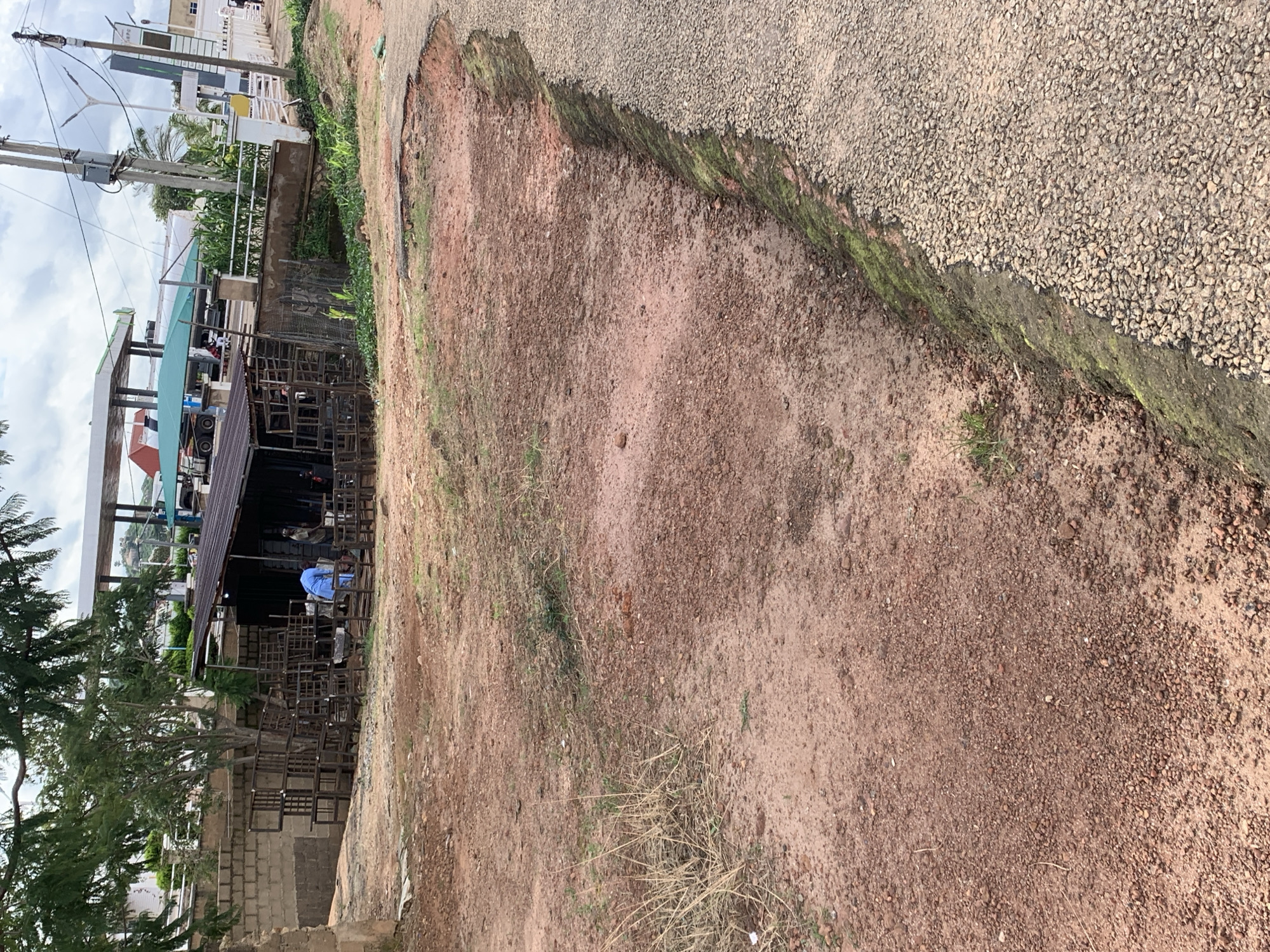 The image shows an outdoor scene in what appears to be a developing area. In the foreground, there is a dirt ground with some dry grass and small stones. To the right, a paved road is visible, with a distinct erosion channel cutting into the embankment between the dirt and the road. The erosion has created a steep, moss-covered edge along the road.
In the middle ground, there is a structure with a corrugated metal roof, resembling a workshop or market stall. Stacked metal frames, possibly for furniture or construction, are arranged outside the structure. A person wearing a blue garment is standing near the structure, and another person is partially visible. Behind this, a low brick wall and some trees can be seen.
In the background, a large, covered structure with white pillars is visible, appearing to be a gas station or a similar commercial establishment. There are also trucks and other vehicles under the canopy. The sky is overcast with some clouds. The overall impression is a scene of everyday activity in an area that may be undergoing construction or development.