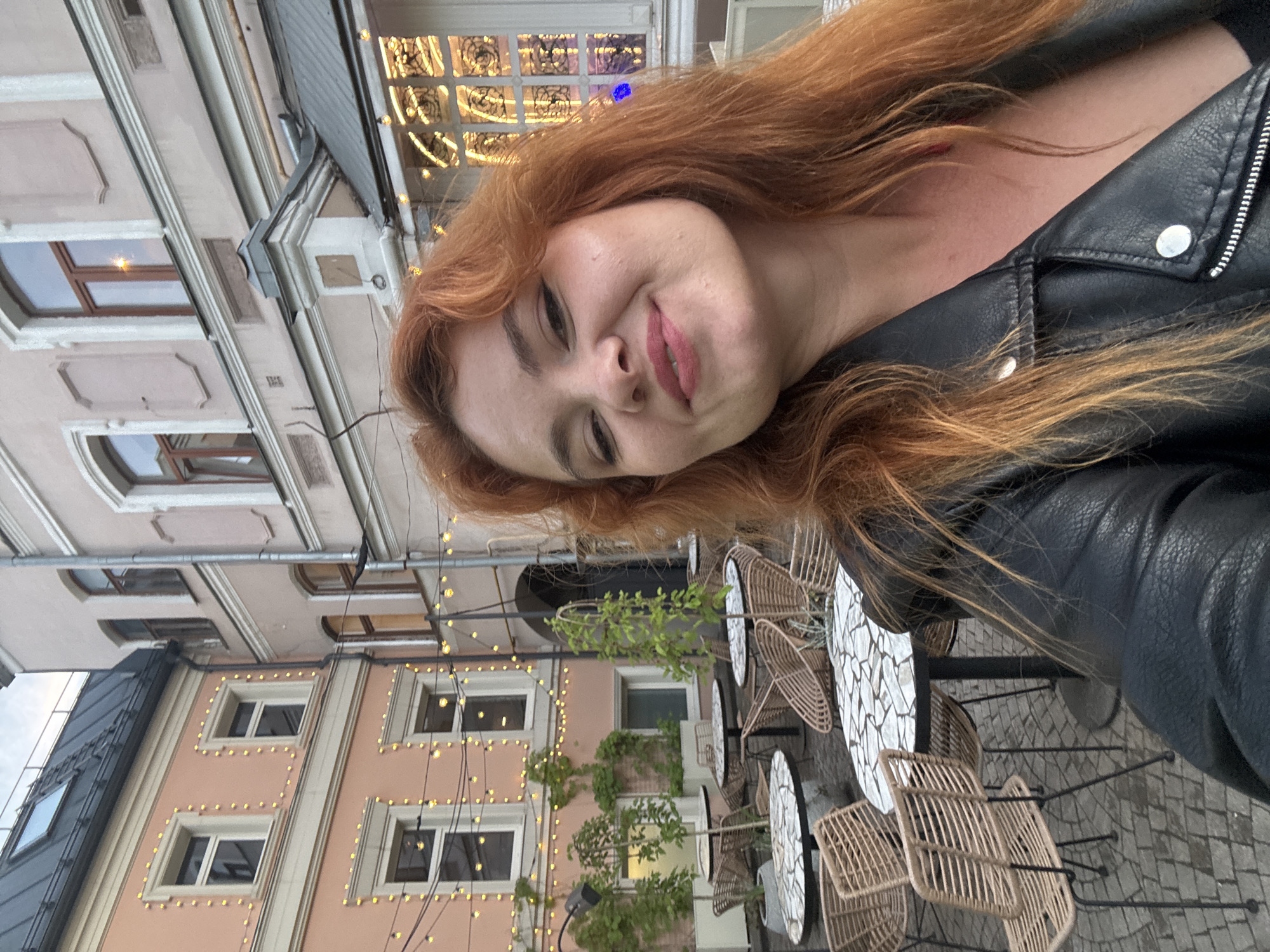 A woman with red hair is taking a selfie in an outdoor cafe in Nizhny Novgorod, Russia. She is wearing a black leather jacket. The cafe has several round tables with mosaic tops and wicker chairs. The background shows buildings decorated with string lights.