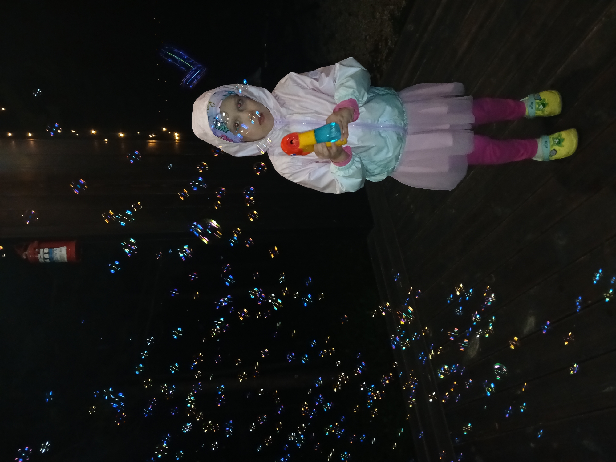 The media file shows a young child playing with a bubble gun at night. The child, wearing a light pink hooded jacket, a light pink tutu, and pink leggings, holds a yellow and blue bubble gun. They are surrounded by numerous iridescent bubbles, reflecting the surrounding lights. In the background, there are blurry lights, suggesting a festive or outdoor setting. A red fire extinguisher is visible on the left side of the frame. The floor appears to be made of dark wooden planks. The time of day is clearly night, and the mood is playful and joyful as the child engages with the bubbles. No visible text is present on the bubble gun or clothing. The scene takes place in Verkhneye Dubrovo, Russia.