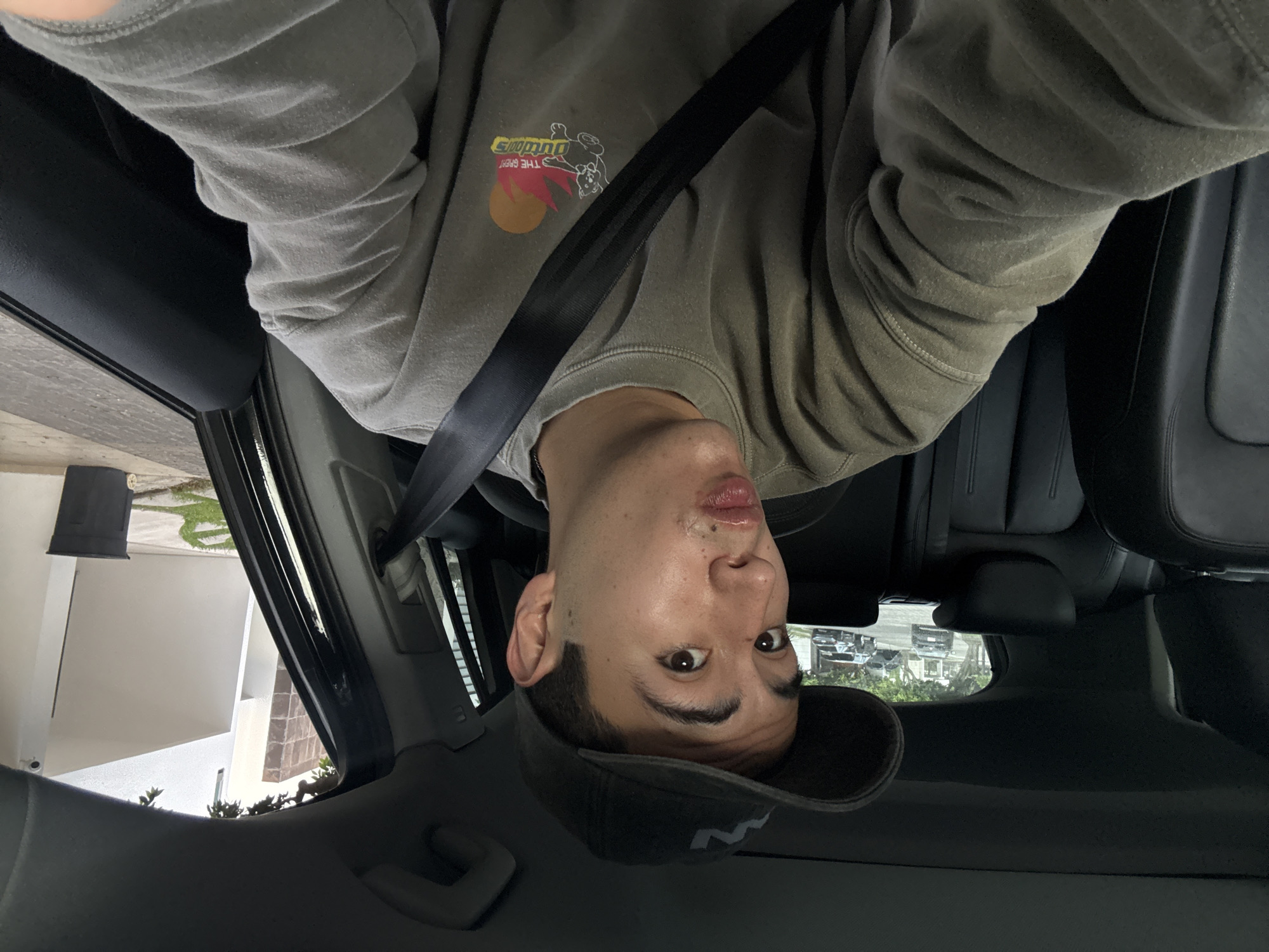 A man is taking a selfie inside a car. He is wearing a gray sweatshirt with a graphic on the chest and a dark-colored baseball cap. He has a seatbelt on. Outside the car window, there is a glimpse of a building with a pot on a wall and some vegetation. Based on the presence of the Spanish language, as per request, it can be inferred that this photo was taken in San Bernardino Tlaxcalancingo, Mexico.