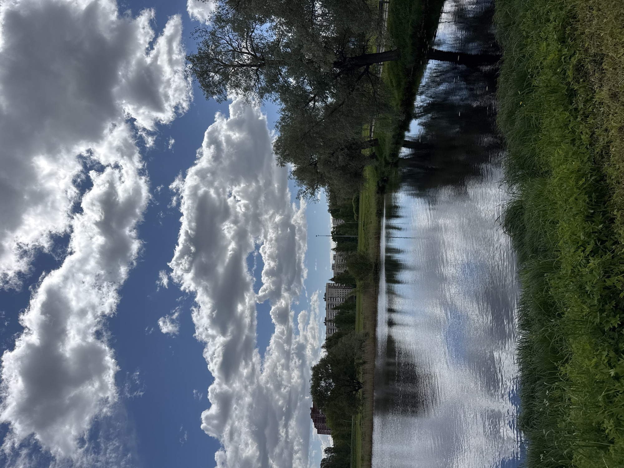 A daytime, outdoor scene depicts a canal reflecting the sky and landscape. The image is taken from a low angle, with the perspective aimed upwards toward the sky and clouds. Fluffy white clouds are scattered across a blue sky, dominating the left side of the image.
To the right, a canal runs vertically, reflecting the clouds, trees, and distant buildings. Trees line the canal’s bank, their branches extending over the water. Green grass grows along the edge of the canal. Further in the distance, buildings are visible, suggesting an urban setting.
The location appears to be a canal in Saint Petersburg, Russia, based on the urban landscape. No people are visible in the shot. The canal's water surface reflects the scene, creating a mirrored effect.