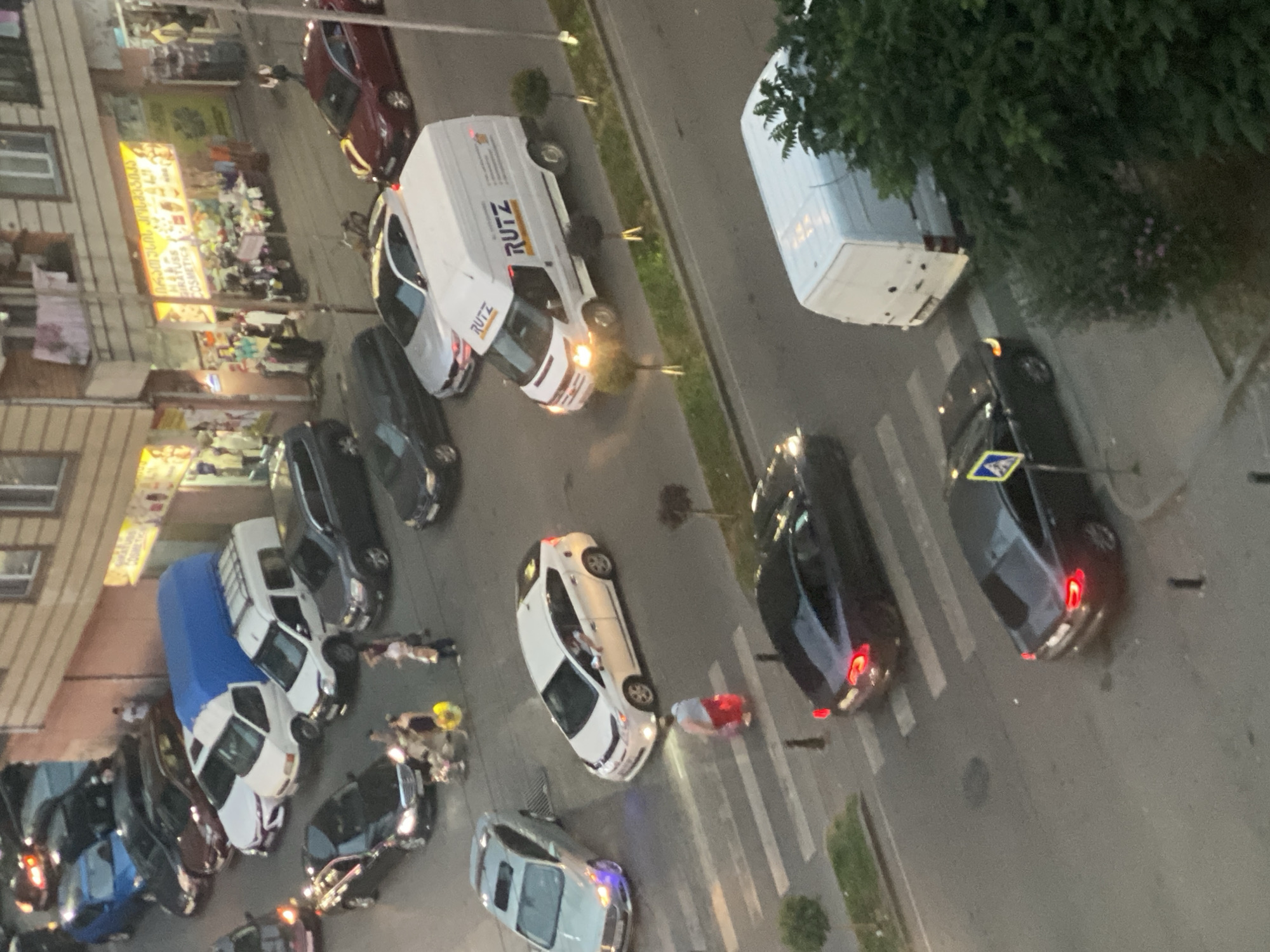 The image shows an aerial view of a street in an urban environment. Several vehicles are present, including cars, vans, and what appears to be a delivery truck. One white van with "RUTZ" branding is in the center right of the frame. A white hybrid sedan is stopped at a crosswalk, and a person in red pants is walking across the crosswalk. Two dark sedans are on the other side of the crosswalk. In the background, there are buildings with storefronts, one of which displays signage in what appears to be Georgian script and advertises cosmetics. Several people are visible on the sidewalk and near the vehicles. The lighting suggests it is either late afternoon or early evening.