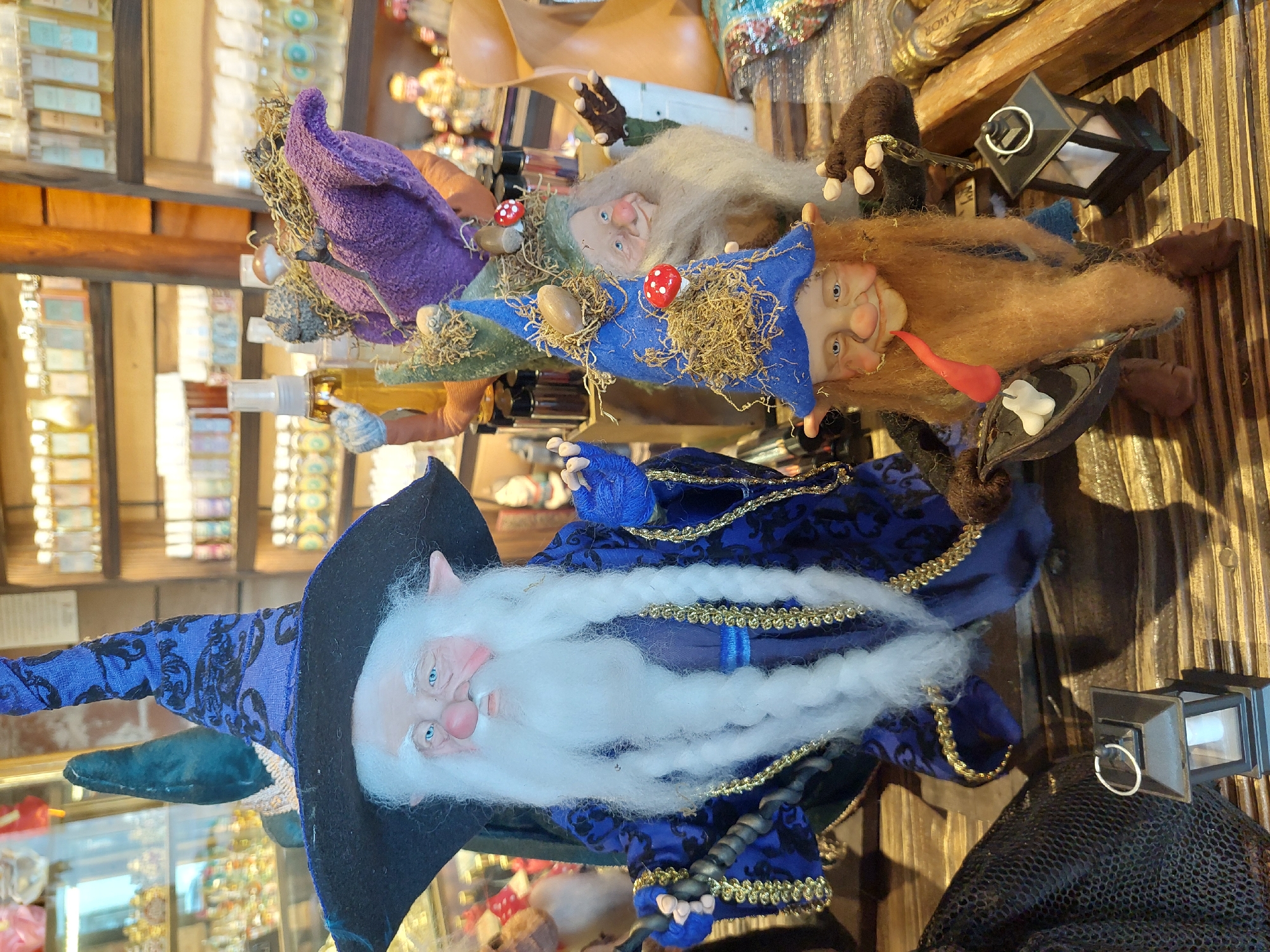 A high-angle, indoor shot depicts several handmade dolls resembling elves or wizards. The most prominent doll is dressed in a dark blue velvet robe with gold trim and a tall, black pointed hat. It has a long, white beard and fair skin with blue eyes. Above it are three smaller, horizontally-aligned dolls. One has a purple hat, another a blue hat, and the third a light brown beard. Each of the three smaller dolls has a red mushroom ornament on its hat. These dolls are displayed on a wooden surface. In the blurred background are shelves filled with bottles and other merchandise, suggesting a retail setting, likely a shop in Buenos Aires, Argentina.