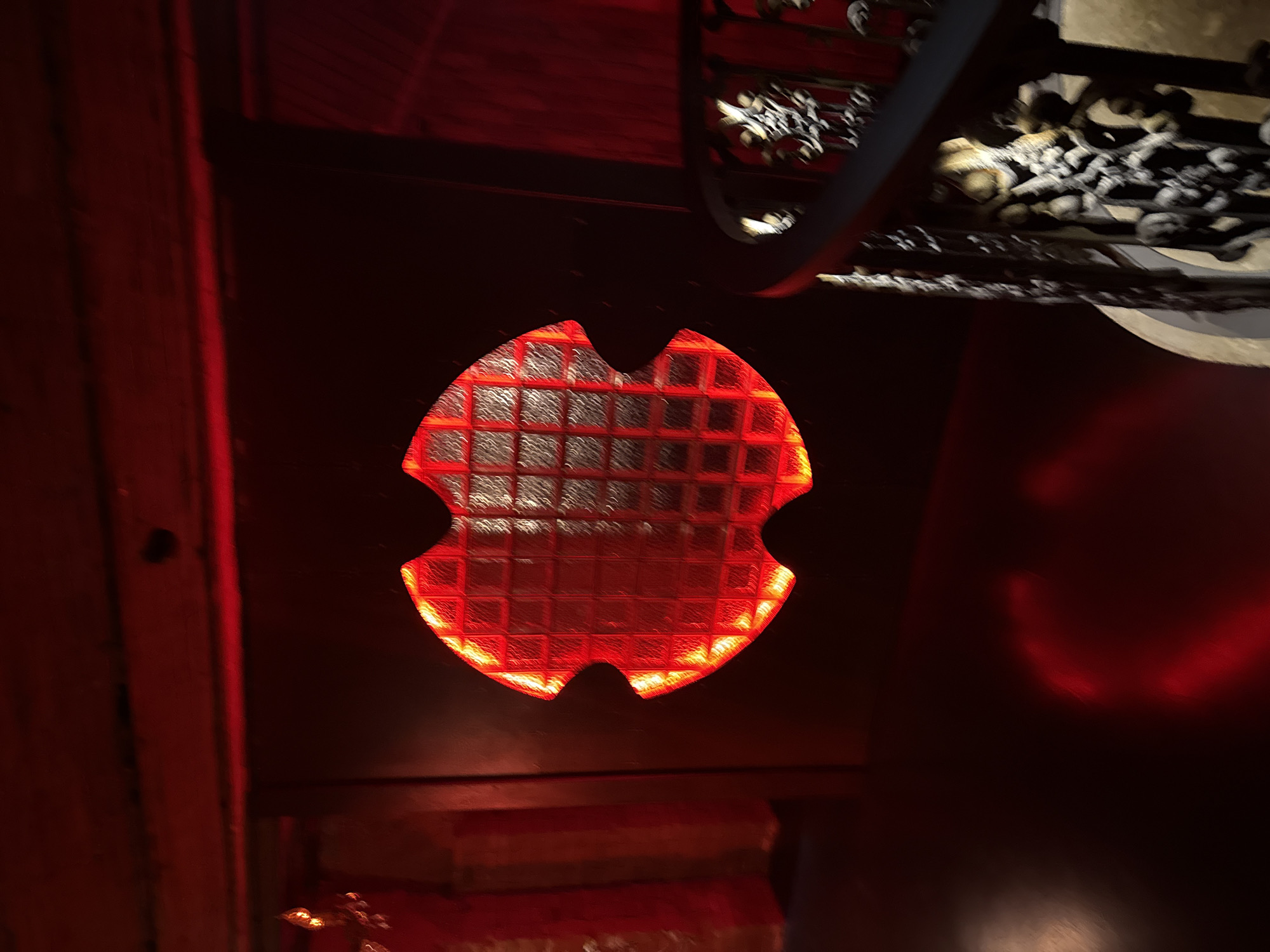 The image depicts a dimly lit interior with a prominent red glow emanating from a uniquely shaped window or illuminated feature in the center. This feature is textured with a grid pattern, resembling glass blocks. To the right, a dark, ornate railing suggests a staircase. The overall ambiance is dark, with red light casting shadows and reflections on the floor and walls. The location is specified as Khamkhi, Russia. No people or specific actions are visible.