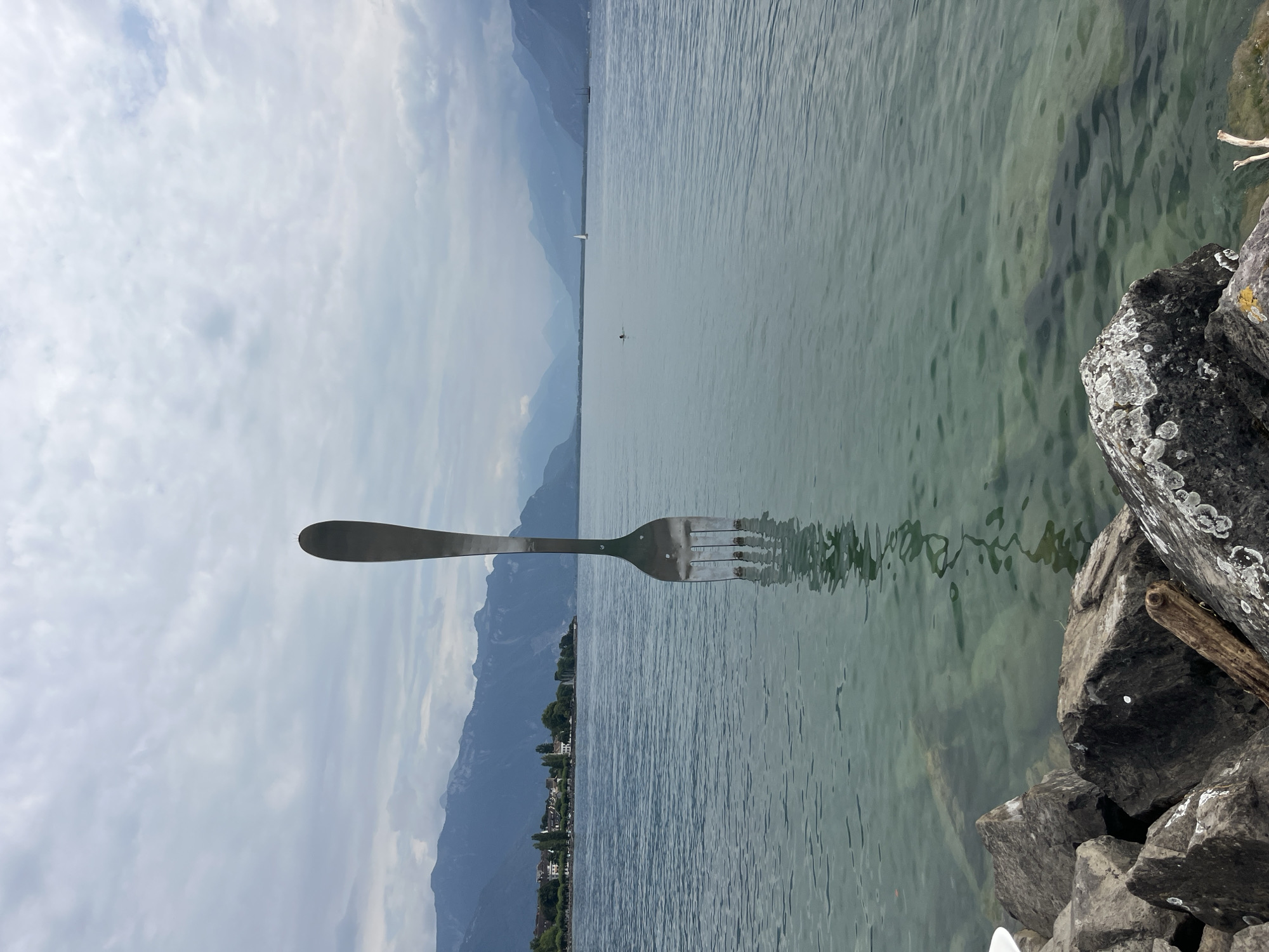 A large, polished stainless steel fork sculpture stands partially submerged in the calm, clear waters of Lake Geneva, in Vevey, Switzerland. The fork's tines are visible just above the waterline, anchoring it in the lakebed. The sculpture is centrally located in the frame, with its reflection clearly visible on the water's surface. In the background, the shoreline of Vevey is visible, featuring buildings, trees, and a distant mountain range under a cloudy sky. Several sailboats and a person swimming are present on the lake, adding to the serene, open-air atmosphere. The foreground features rough, grey rocks lining the edge of the lake, with some driftwood visible amongst them.