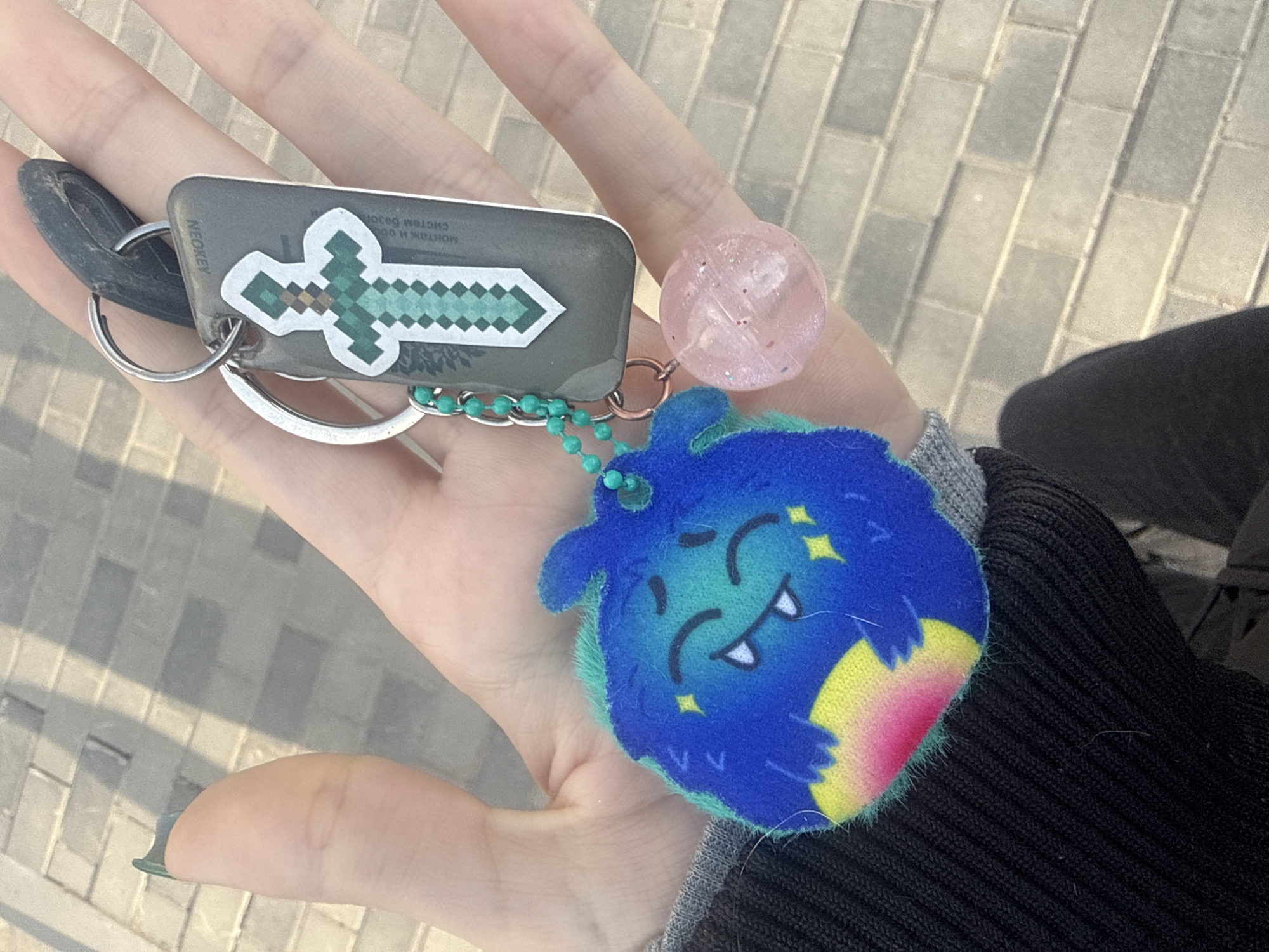A first-person perspective photograph shows a left hand holding a keychain with various trinkets. The keychain holds a car key with a "NEOKEY" label, adorned with a sticker of a pixelated Minecraft sword. Additionally, there's a pink glittery ball and a plush blue monster charm featuring a crescent moon and fangs. The hand appears to be in a palm-up position, displaying the keychain.
The background features a paved area, likely a sidewalk. In the periphery, a portion of a black sweater is visible, suggesting the person holding the keys is wearing it. The setting seems to be outdoors, with the paving stones indicating an urban environment.
There is no definitive evidence in the image to confirm the location. Therefore, the inclusion of the city of Saint Petersburg, Russia, cannot be verified based on the provided image alone.