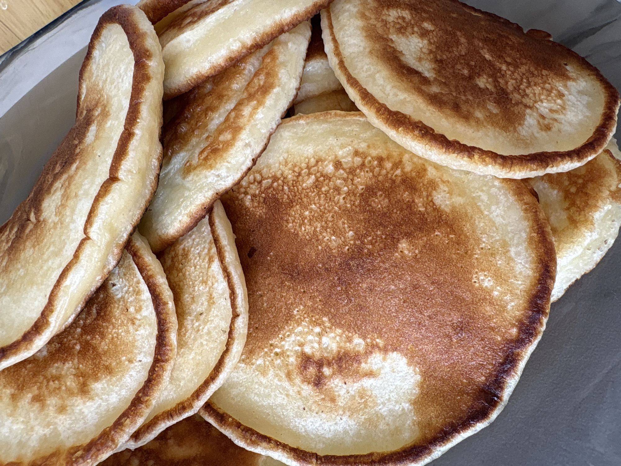 A close-up, overhead shot captures a stack of golden-brown pancakes in a bowl. The pancakes have a slightly uneven, rustic texture with visible bubbles on their surface, indicating they were recently cooked. Their edges are a darker brown, creating a nice contrast with the lighter, golden-yellow interior. The stack is piled high, with some pancakes overlapping others, creating a sense of abundance. The lighting suggests it could be daytime, with the natural light highlighting the warm tones of the pancakes. The background is out of focus, but appears to be a neutral surface, possibly a table or countertop. There are no people visible, and the scene focuses solely on the appetizing presentation of the pancakes. The location context of Засечное, Russia, is not visually evident within the image itself, as the scene is domestic and lacks specific geographical markers. No text is visible.
