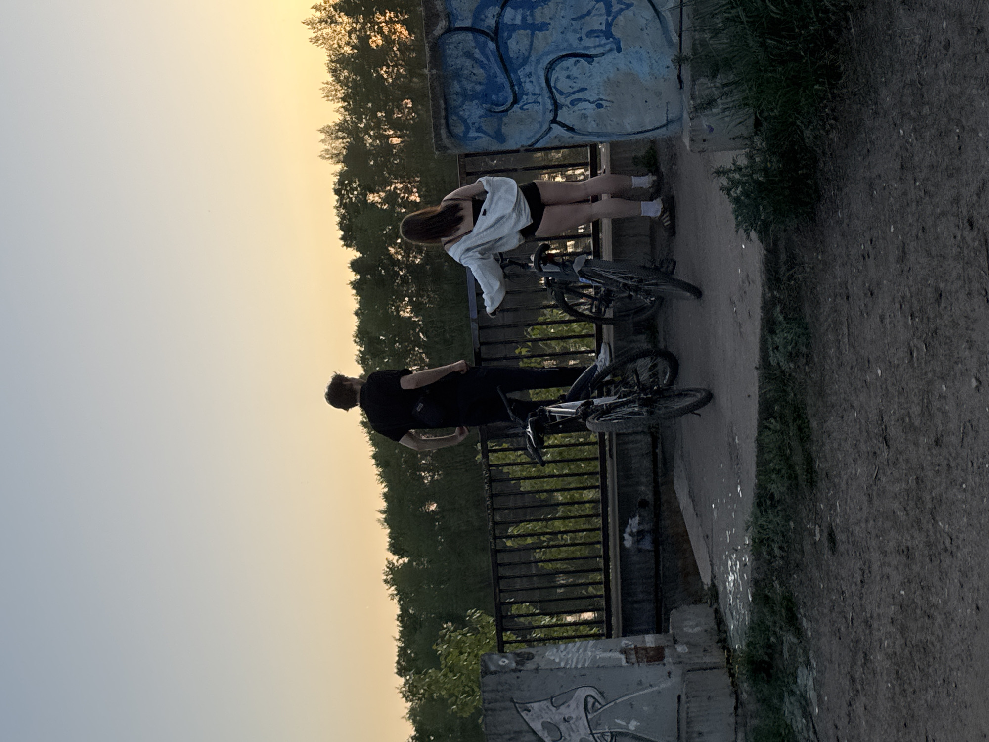 On a bridge in Chaikovsky, Russia, a male and female stand by their bicycles, both facing the bridge railing. The female, dressed in shorts and a white jacket, leans over the railing, while the male, wearing a black shirt and pants, stands next to her. Behind them, trees line the horizon beneath a dusky sky. Graffiti adorns the concrete structure to the right of the couple and a similar concrete structure is at the bottom of the frame. The ground to the right of the image is dirt, with grass and weeds growing beside the bridge.