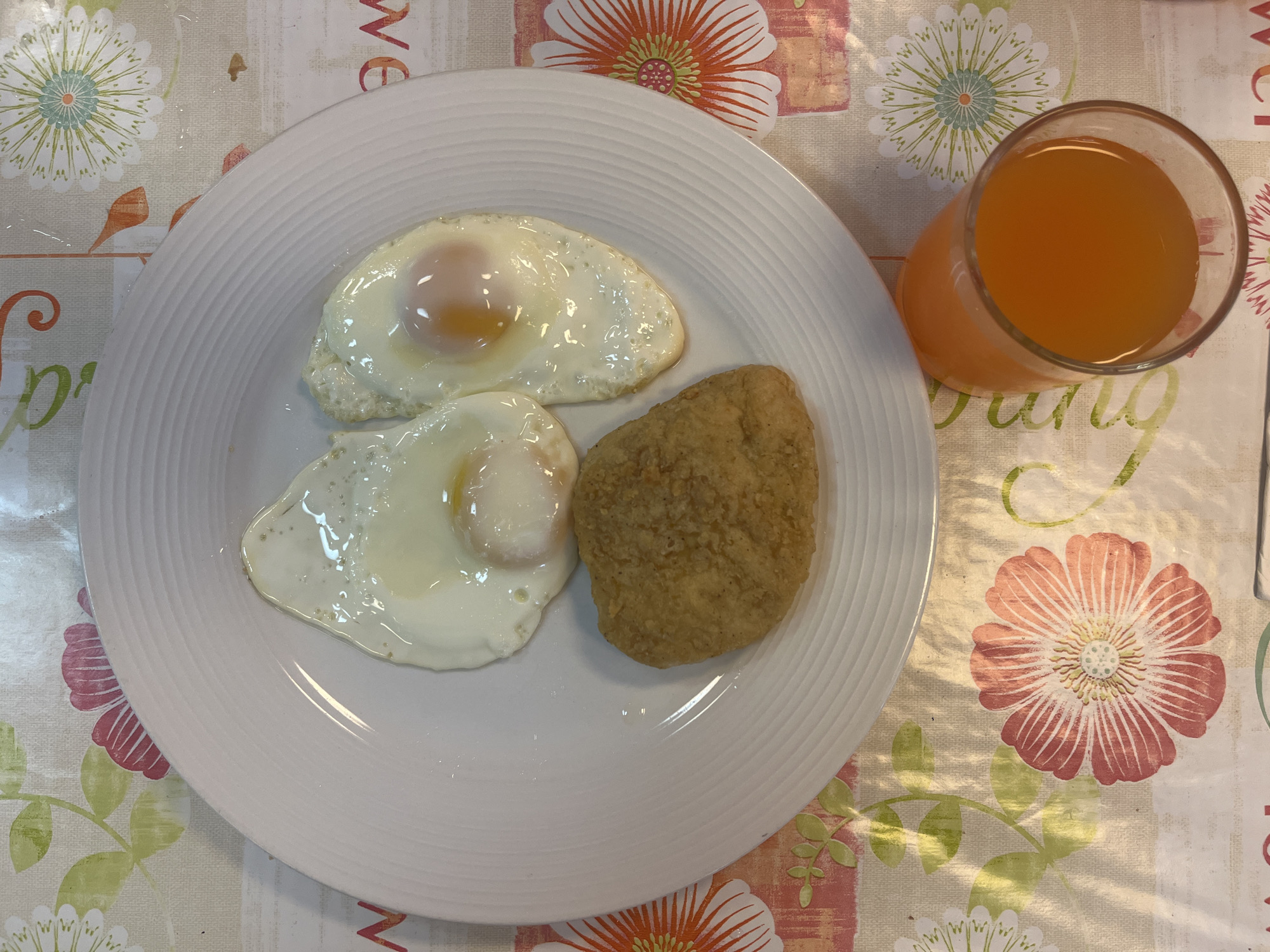 A breakfast plate containing two fried eggs and a hash brown sits next to a glass of orange juice on a floral patterned tablecloth. The tablecloth features large flowers in shades of red, orange, and white, along with smaller green leaves. The plate is white with concentric rings near the edge. The fried eggs have runny yolks and slightly browned edges. The hash brown is golden brown and appears crispy. The orange juice is a vibrant orange color and fills the glass almost to the top. The location is Unknown, Unknown.