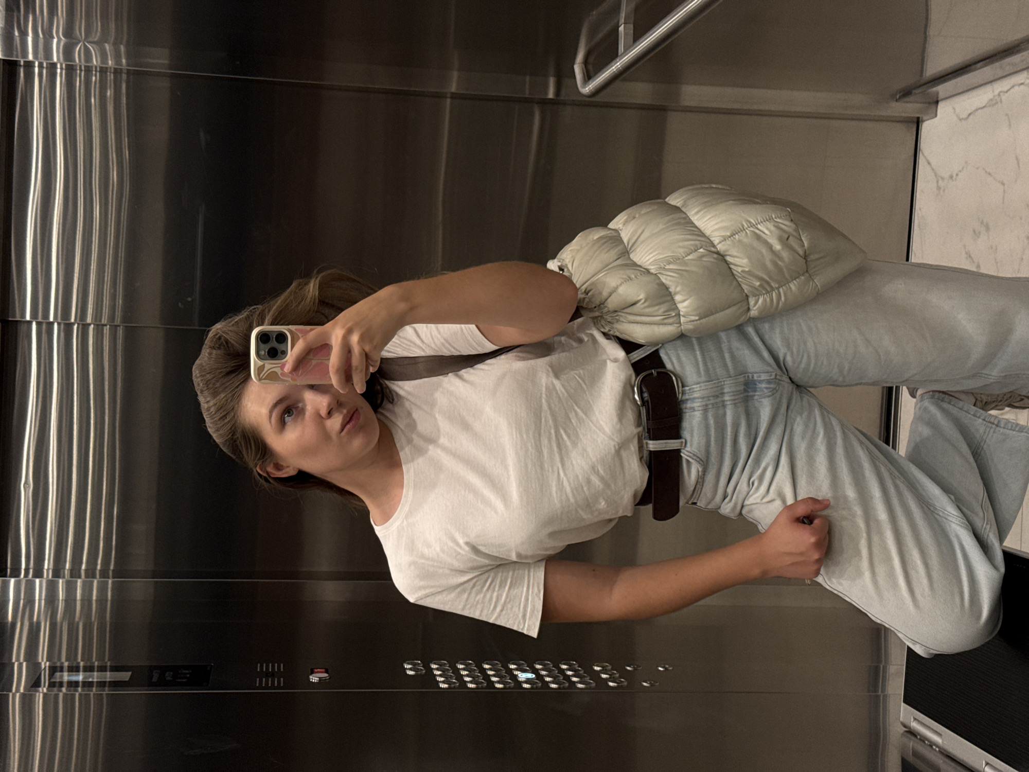 A woman is taking a mirror selfie in what appears to be an elevator. She is wearing a white t-shirt, light-wash jeans, and a brown belt. She has a light-colored puffer bag slung over her shoulder. Her hair is pulled back. The elevator walls are made of brushed stainless steel, with a handrail visible at the top. The elevator buttons are also visible on the wall. The scene is dimly lit. The user's phone, with a pink and beige case, is visible as she holds it to take the picture. The overall mood seems casual and perhaps a bit playful, given the selfie. There is no visible text in the image.