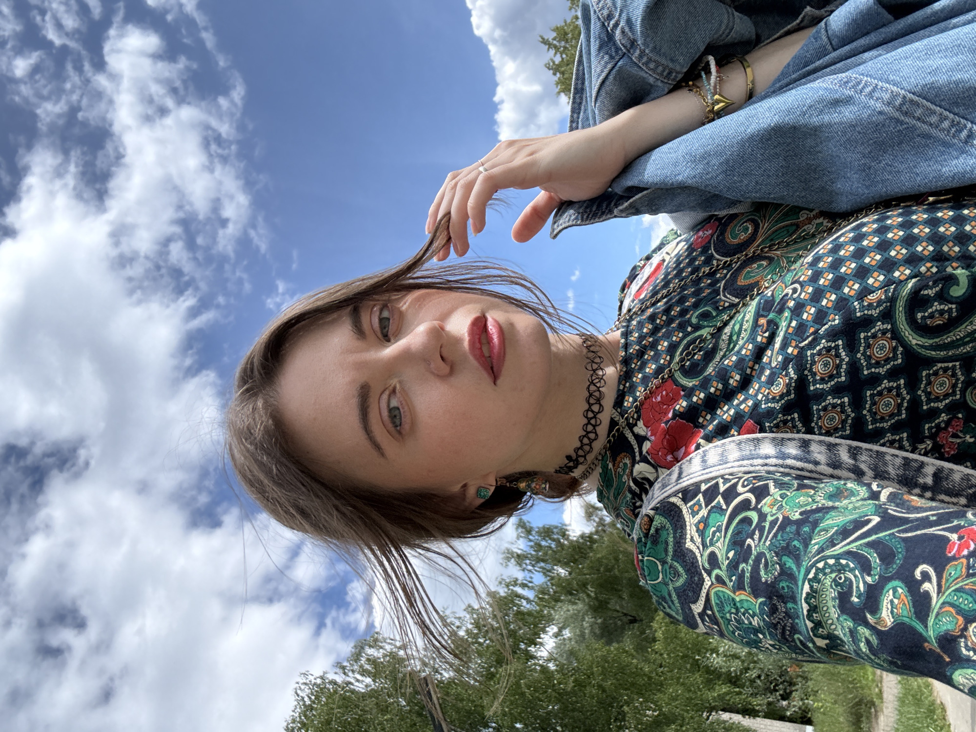 A young woman is captured in a low-angle, sideways selfie. She is looking directly at the camera with a neutral expression, her gaze intense. Her right hand is raised, lightly touching her hair, while her left arm is bent and resting against her body. She is wearing a dark, patterned top with a bohemian-inspired design featuring red flowers and intricate paisley-like motifs. A denim jacket is draped over her shoulders, and she accessorizes with a delicate silver ring on her right hand, a black choker necklace, and dangling earrings. On her left wrist, she sports several bracelets, including one with a gold heart charm.
The background is a vibrant blue sky filled with fluffy white clouds, suggesting it is daytime. Lush green trees are visible in the lower portion of the frame, hinting at an outdoor setting, possibly a park or a garden. The lighting indicates it is a bright, sunny day. The overall mood is relaxed and natural, with the woman appearing comfortable in her surroundings. There is no visible text in the image.