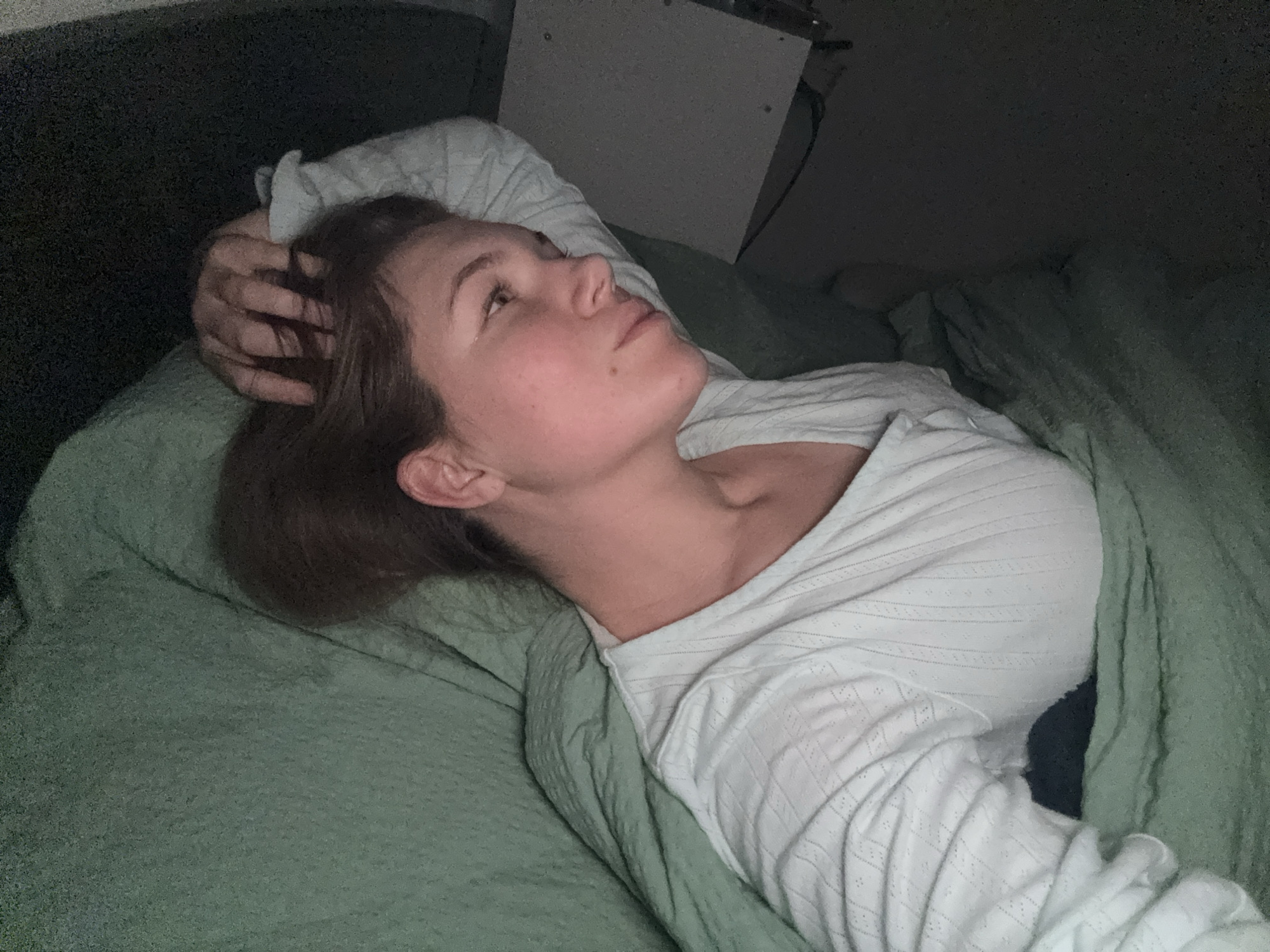 A woman lies in bed, her head resting on a pillow, her right hand touching her hair. She is looking upwards. She is covered with a green duvet, and is wearing a white patterned top. Behind her head is a bedside table. The location of this image is not apparent.