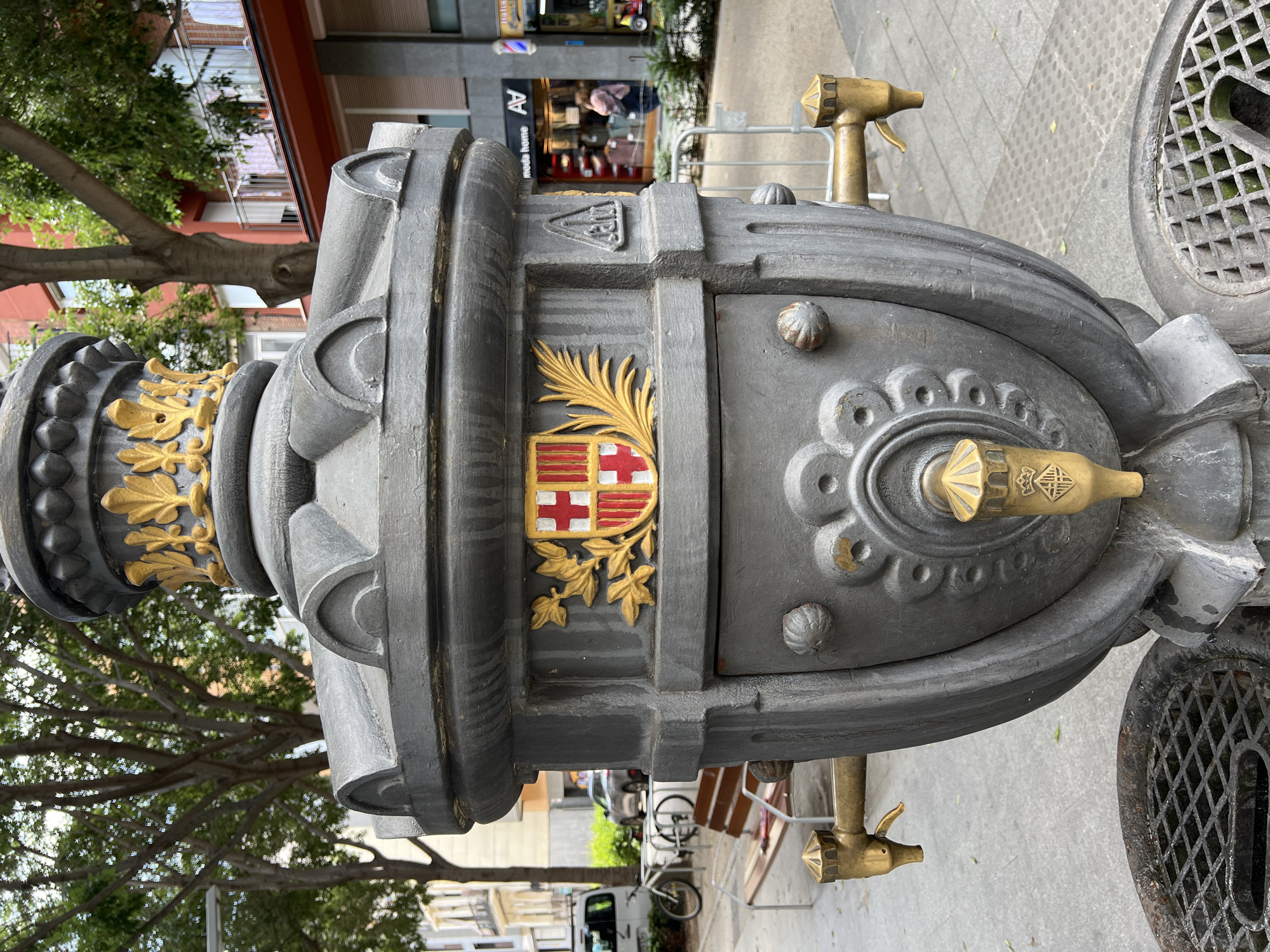 The image shows a close-up, eye-level view of an ornate, dark gray cast-iron public fountain in Barcelona, Spain. The fountain features a prominent coat of arms on its front, displaying a red cross on a white background, accompanied by four red stripes on a yellow background, within a shield. This is the coat of arms of Barcelona. To the sides of the coat of arms are decorative golden foliage. The fountain has two brass spigots on either side, and a central brass faucet with a decorative crown motif. The fountain is positioned on a gray paved area, with a metal grate visible at the bottom foreground. In the background, trees with green foliage line the street, and buildings with red and white facades are visible, along with shops and a balcony with laundry hanging out. The overall scene suggests an urban outdoor setting in a European city.