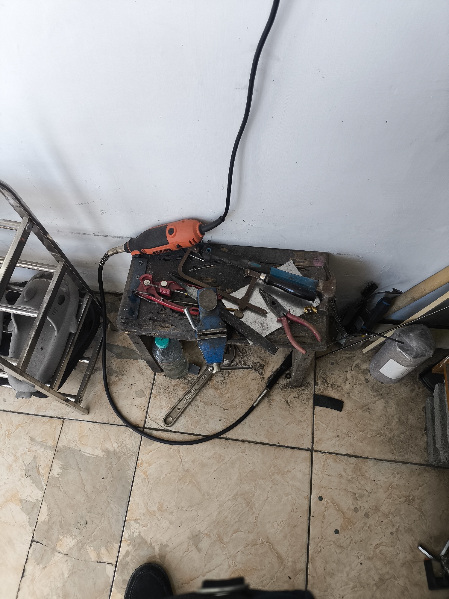 An overhead shot captures a cluttered workbench and surrounding area on a tiled floor, likely in Lumajang, Indonesia. The workbench itself is a rustic wooden stool holding an array of tools. Prominently featured is an orange and black rotary tool connected to a black cord that snakes upwards and out of frame. Adjacent to it are various pliers, including a pair of red-handled wire cutters, and a blue vise clamped to the stool. A bottle of water sits on the floor beneath the stool. To the left of the workbench, a metal rack holds a gray, helmet-like object. The floor is covered in light-colored marble-patterned tiles. To the right of the workbench, various other items are visible, including what appears to be a bundled object wrapped in plastic and some wooden planks leaning against a wall. The wall itself is painted white and shows signs of some scuff marks. A dark shoe is visible in the bottom of the frame, suggesting a person is present, though no individuals are fully depicted. The overall impression is that of a functional, albeit messy, workspace.
