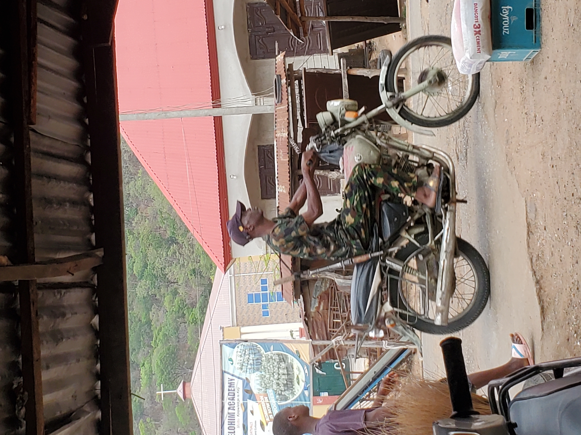 A man wearing a military-style shirt and a baseball cap is seen on a vintage motorcycle parked in what appears to be a street in Mararaba, Nigeria. He seems to be fixing the bike. On the front wheel of the motorcycle is a bag of Dangote cement and a crate of Fayrouz. In the background, there is a building with a sign that reads "Elohim Academy". In the foreground, a child sits on the ground. Other buildings are visible across the street. The overall environment suggests an urban area with commercial or residential buildings.