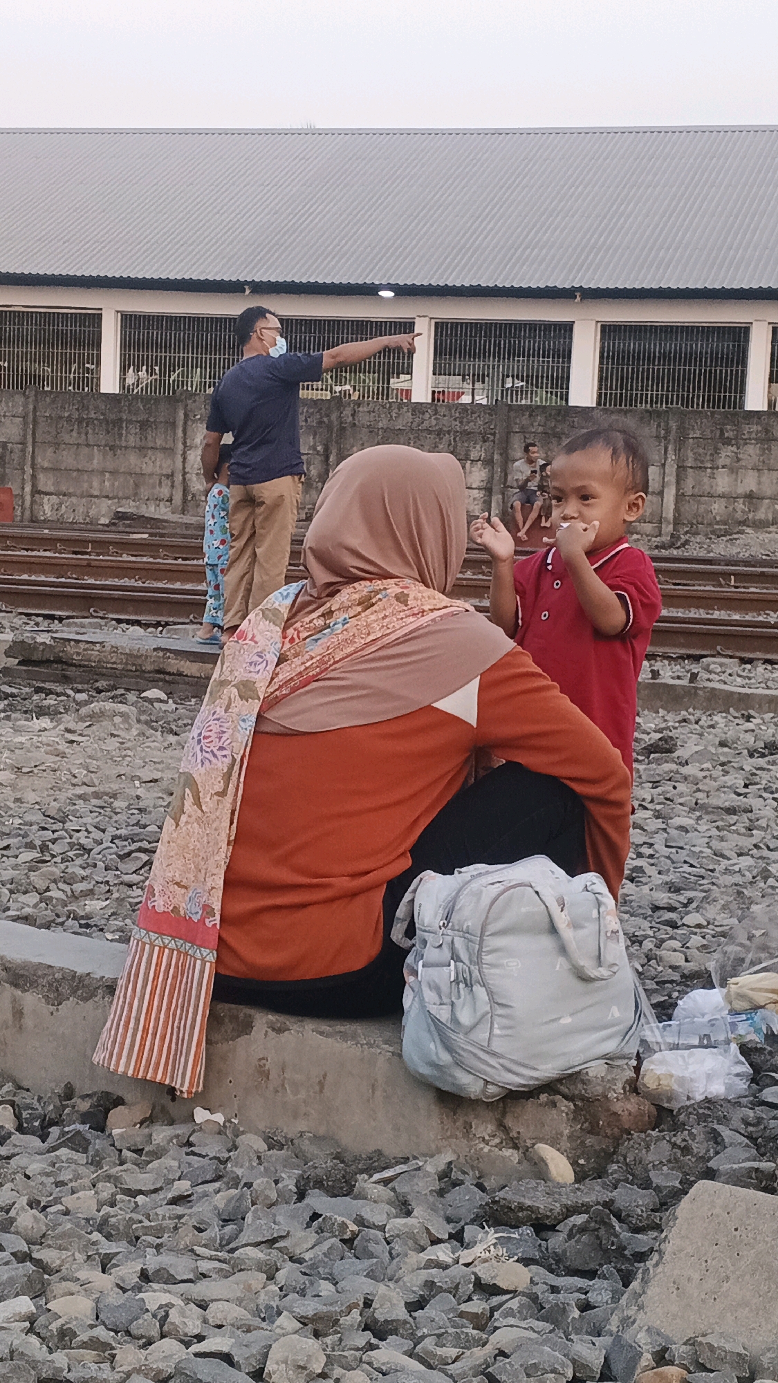 A group of people are near a set of train tracks. A man wearing a blue t-shirt and a mask is pointing to the left. A woman wearing a headscarf and an orange shirt is sitting on a concrete barrier, facing away from the camera. A young child in a red shirt is standing in front of the woman, facing the camera and eating something. A light blue backpack is on the ground next to the woman. In the background, there is a building with a gray roof and barred windows. The ground is covered in gravel and rocks. The scene appears to be outdoors, potentially at a train station in Semarang, Indonesia.